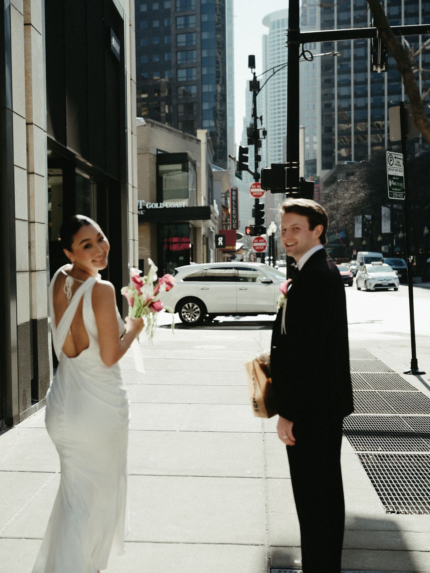Bride and groom smile on a sunny Chicago city sidewalk, bride in white backless gown holding bouquet.
