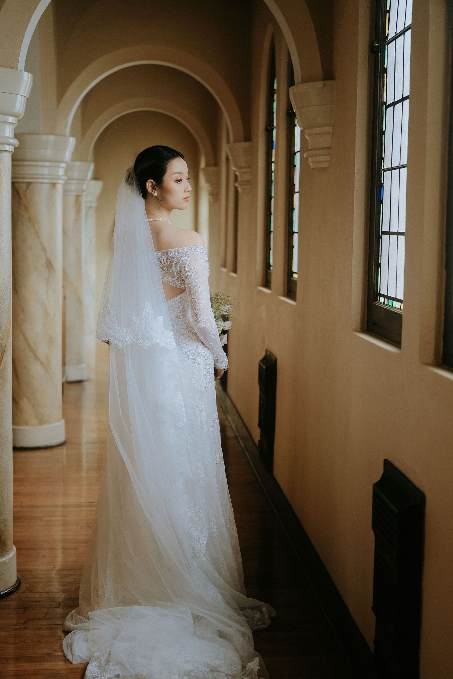 Bride in off-shoulder lace wedding gown with veil stands in sunlit arched hallway of elegant venue.