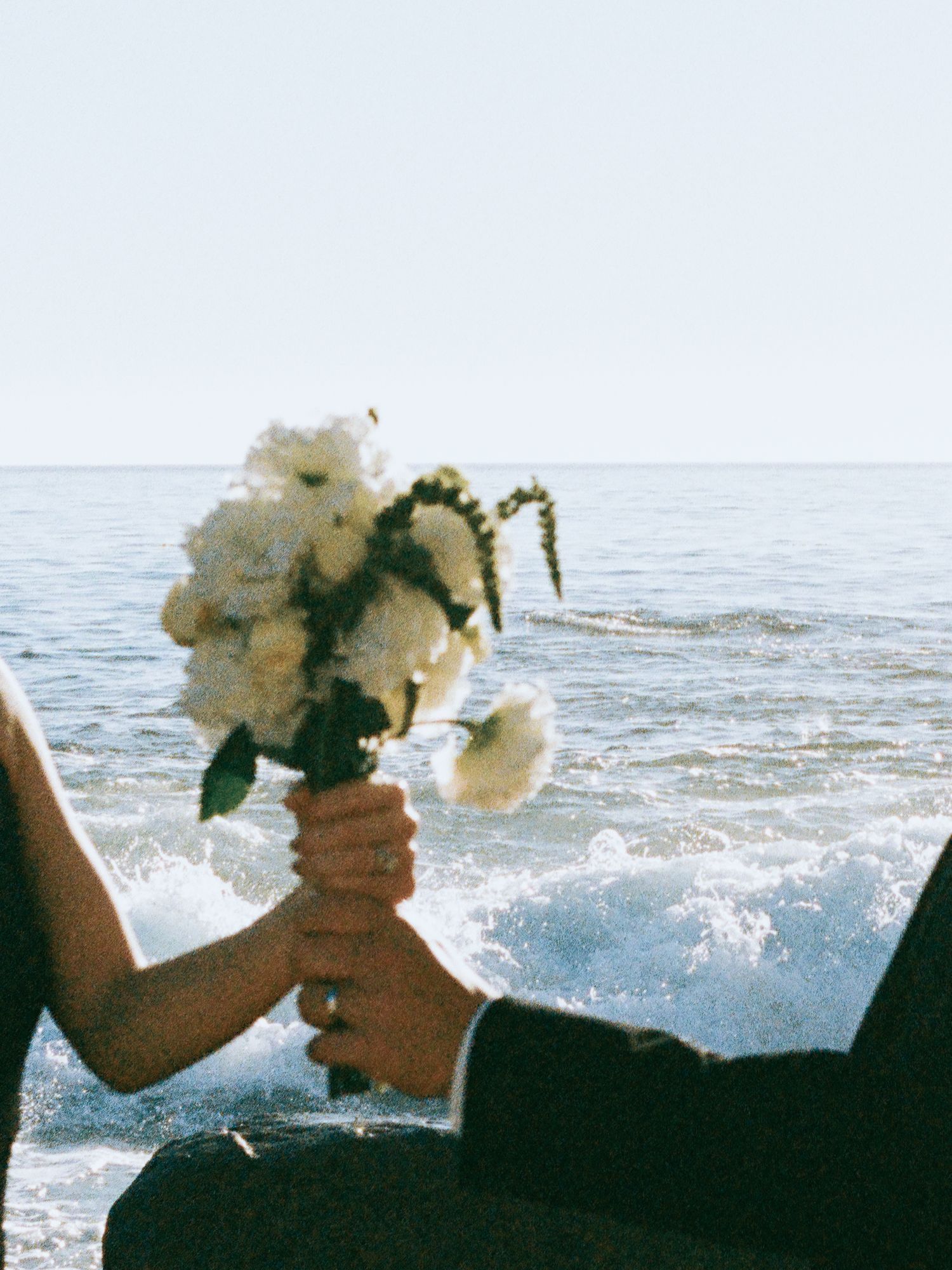 Bride and groom holding white peony bouquet near ocean waves on wedding day.