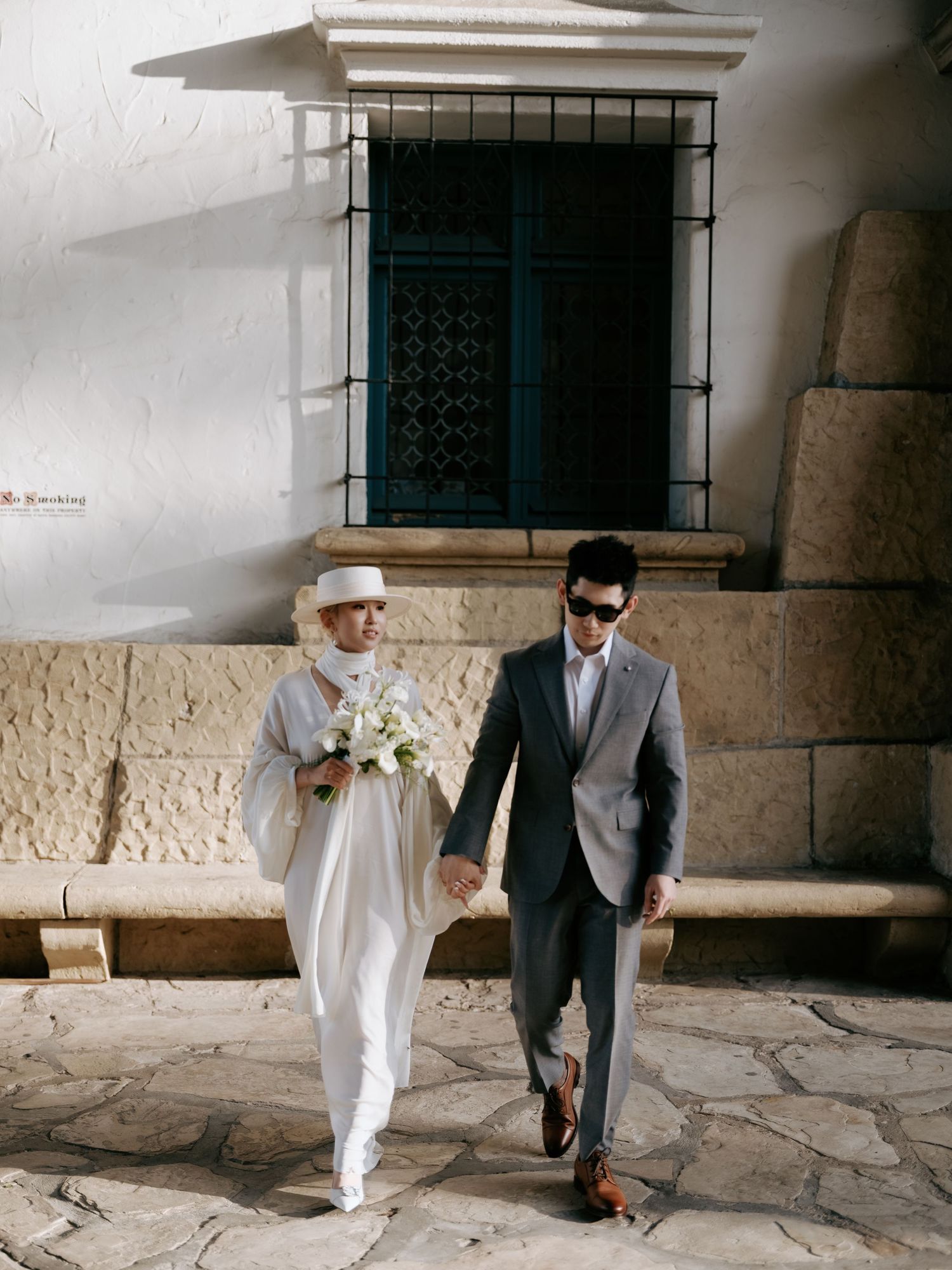Bride and groom walk hand in hand along stone courtyard of a historic venue.