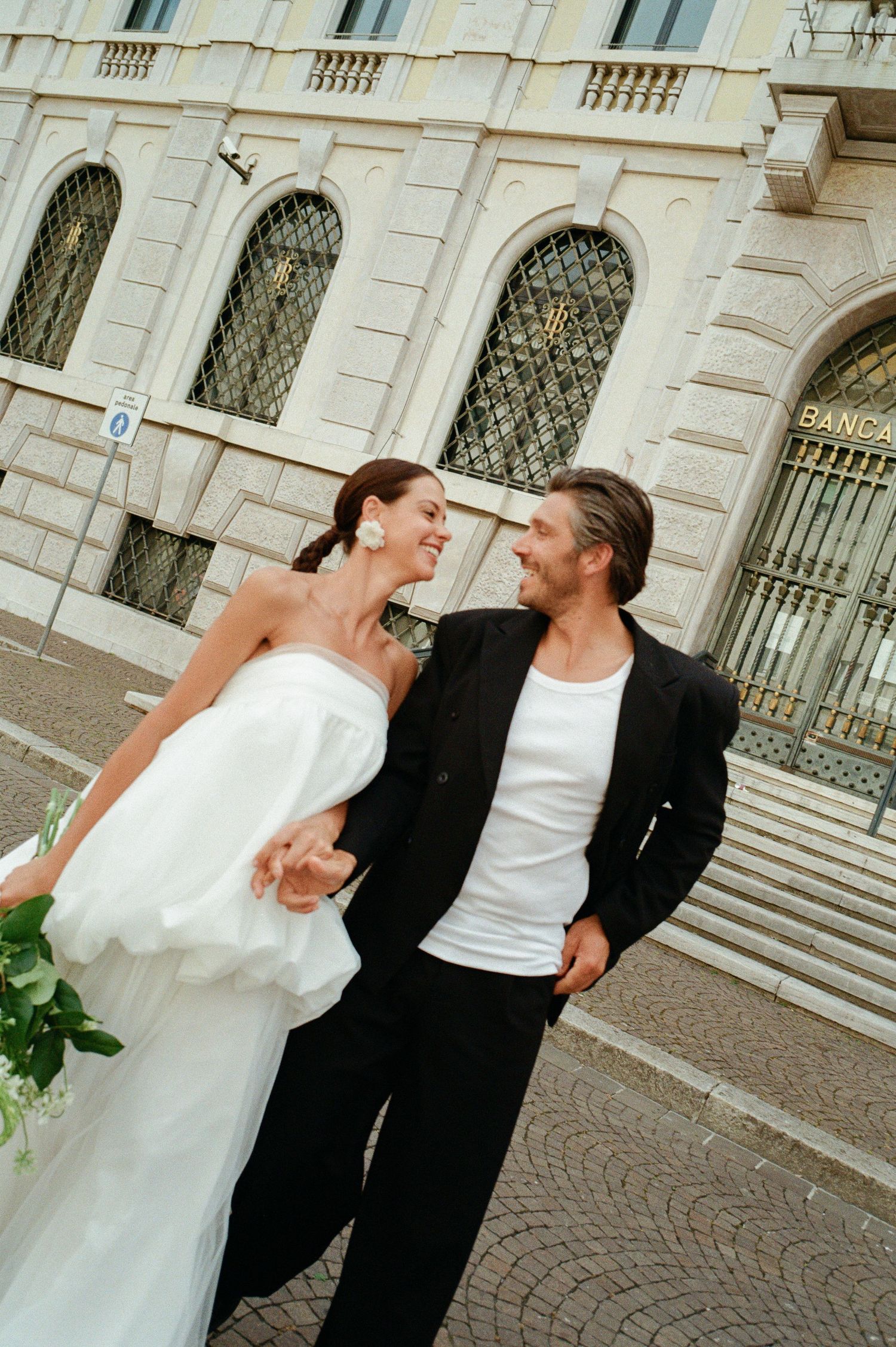 Bride in white strapless gown and groom in black jacket laugh together outside Banca d Italia.