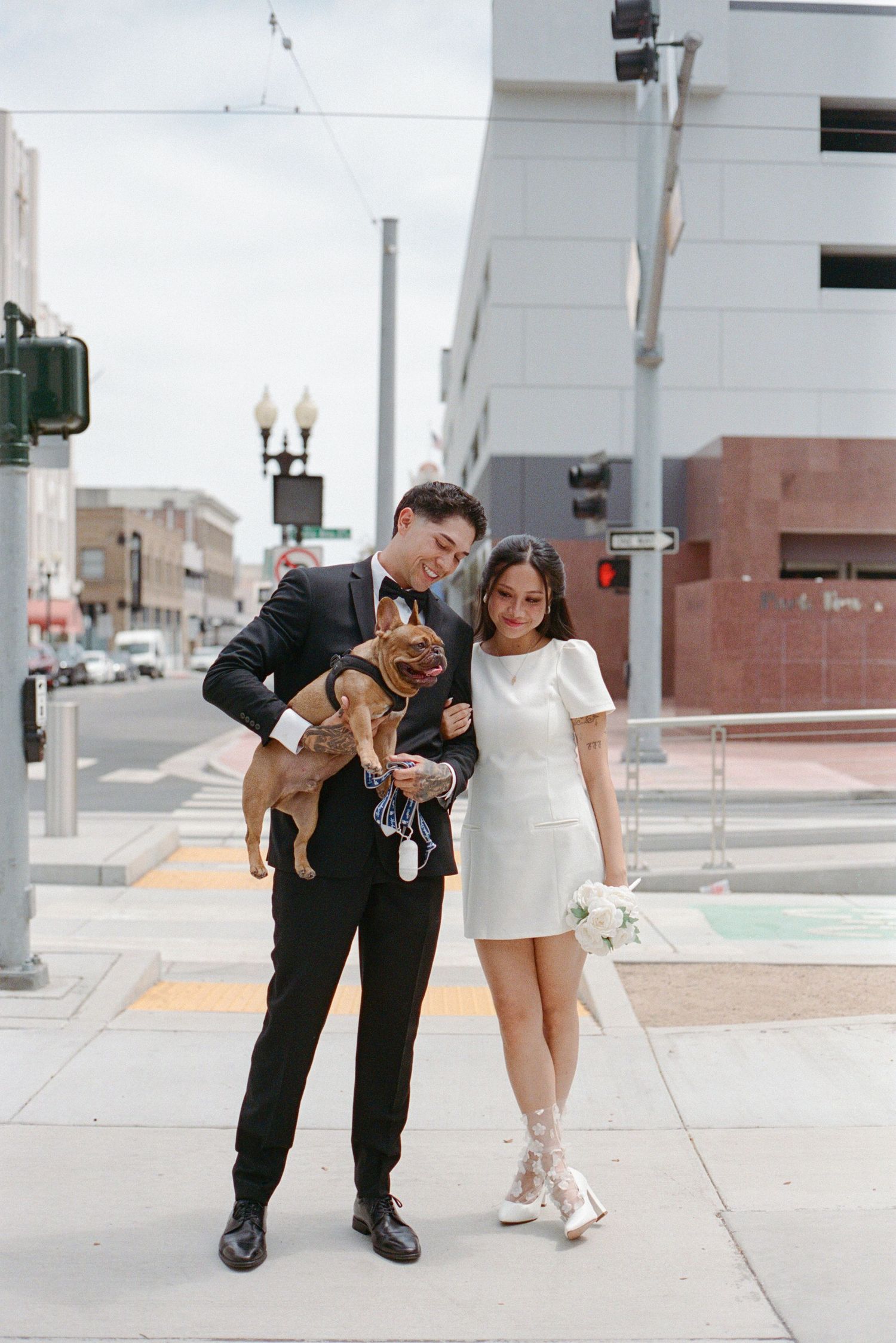 Newlywed couple poses with their French Bulldog on a city sidewalk, bride holding a white bouquet.