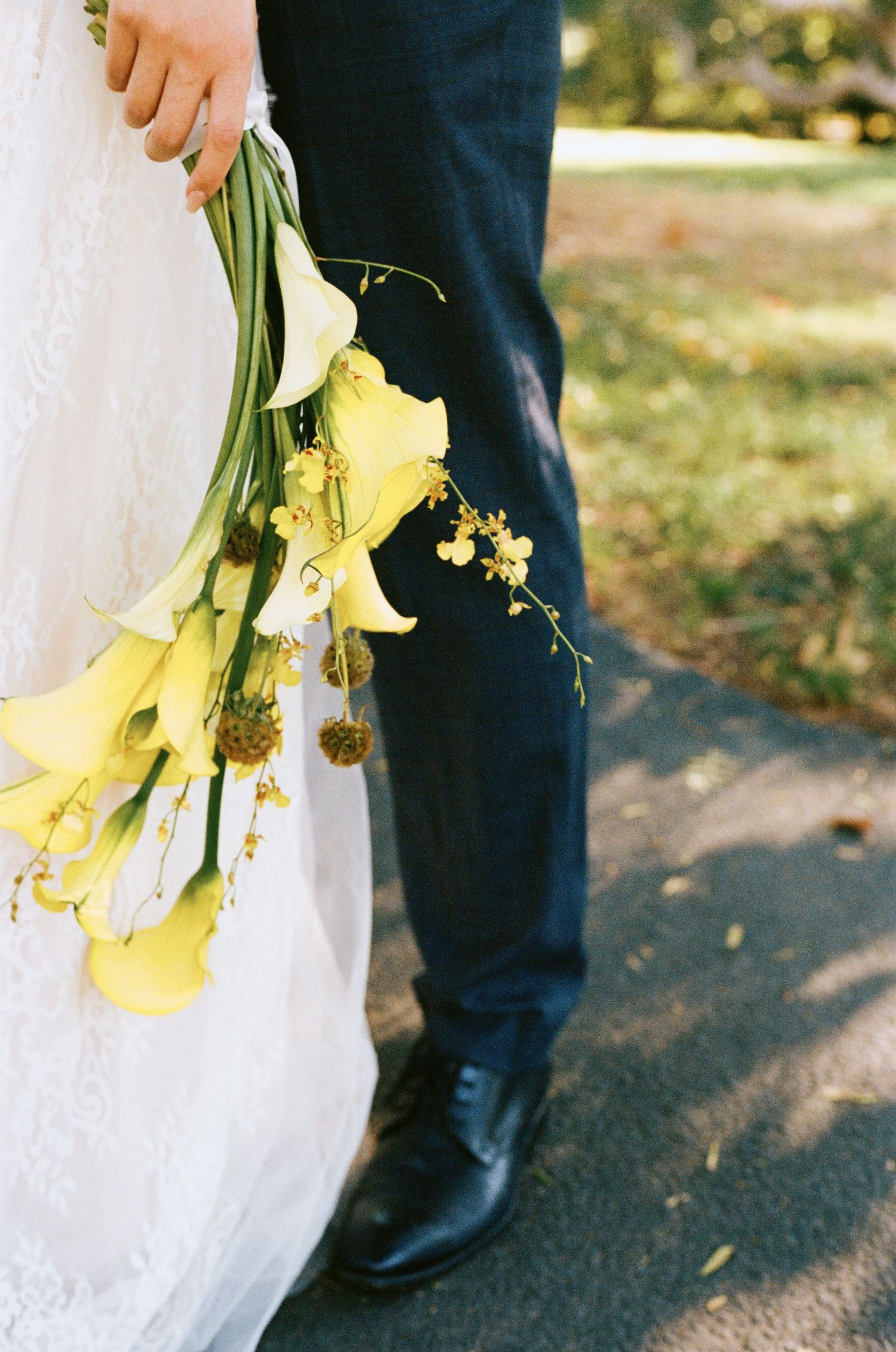 Close-up of yellow calla lily wedding bouquet held by bride in lace gown beside groom in black trousers.