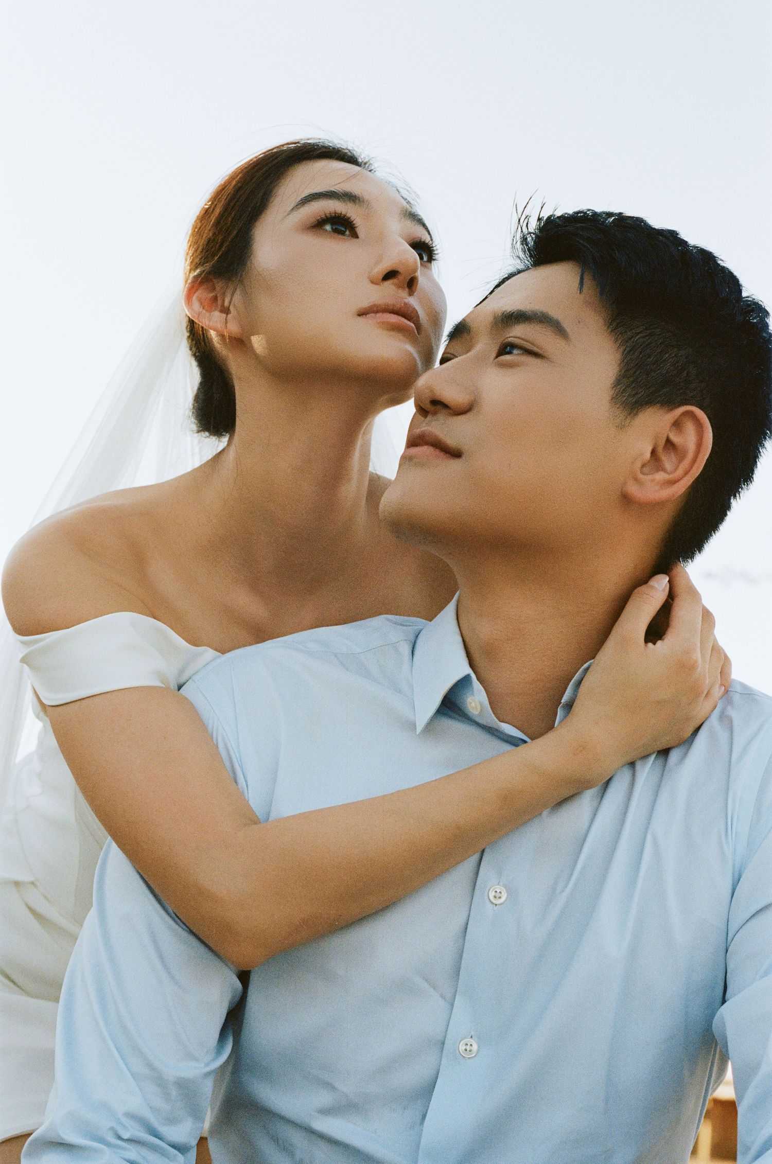 Asian bride in off-shoulder dress and veil embraces groom tenderly under a bright open sky.