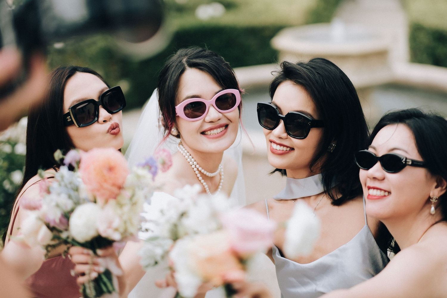 Smiling bride and bridesmaids wearing stylish sunglasses posing together outdoors with pastel wedding bouquets.