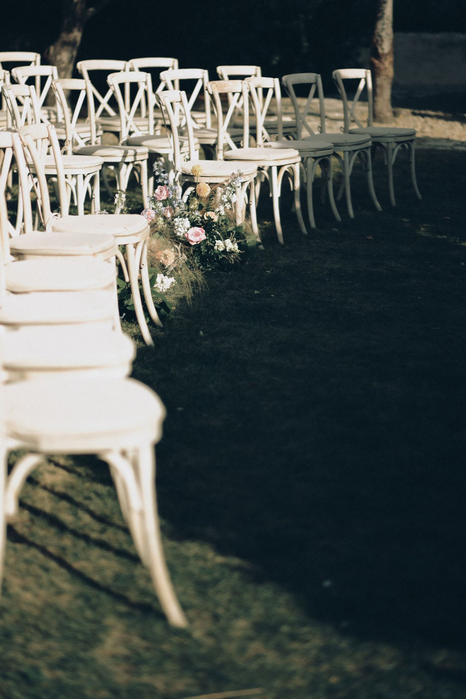 Rows of white crossback chairs lined with floral arrangements on green lawn for an outdoor wedding ceremony.