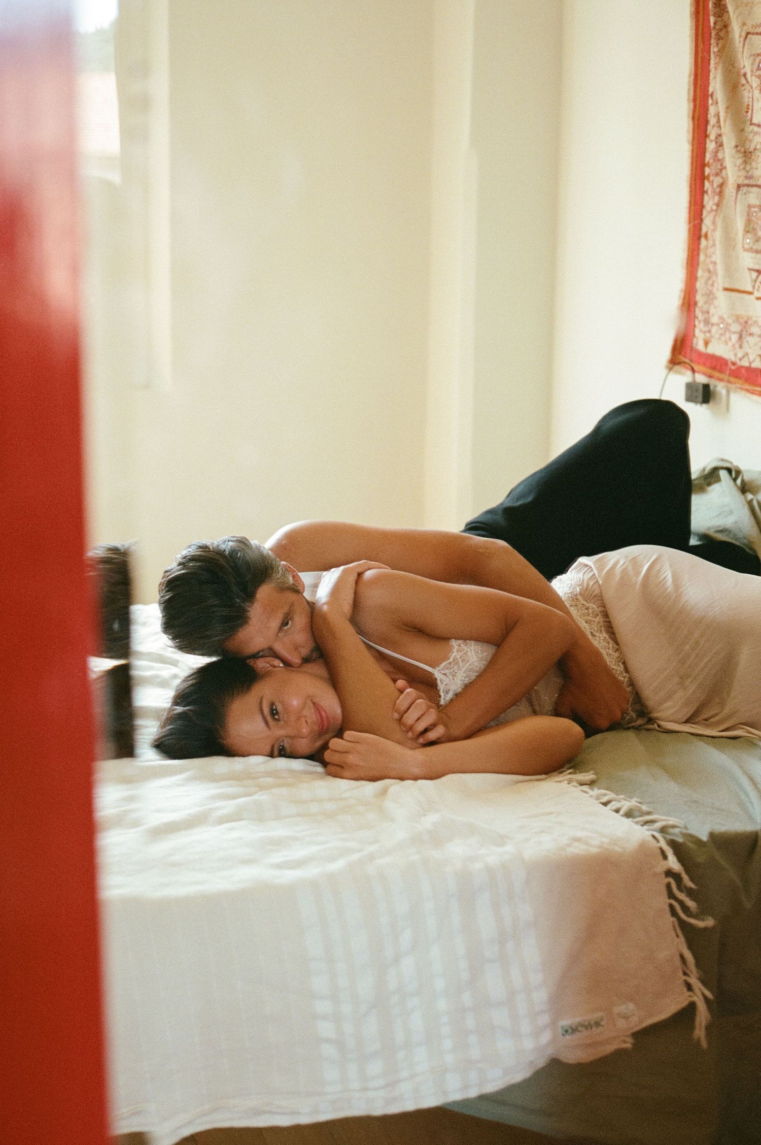 Man tenderly embracing woman from behind as they lie together on a white bed.