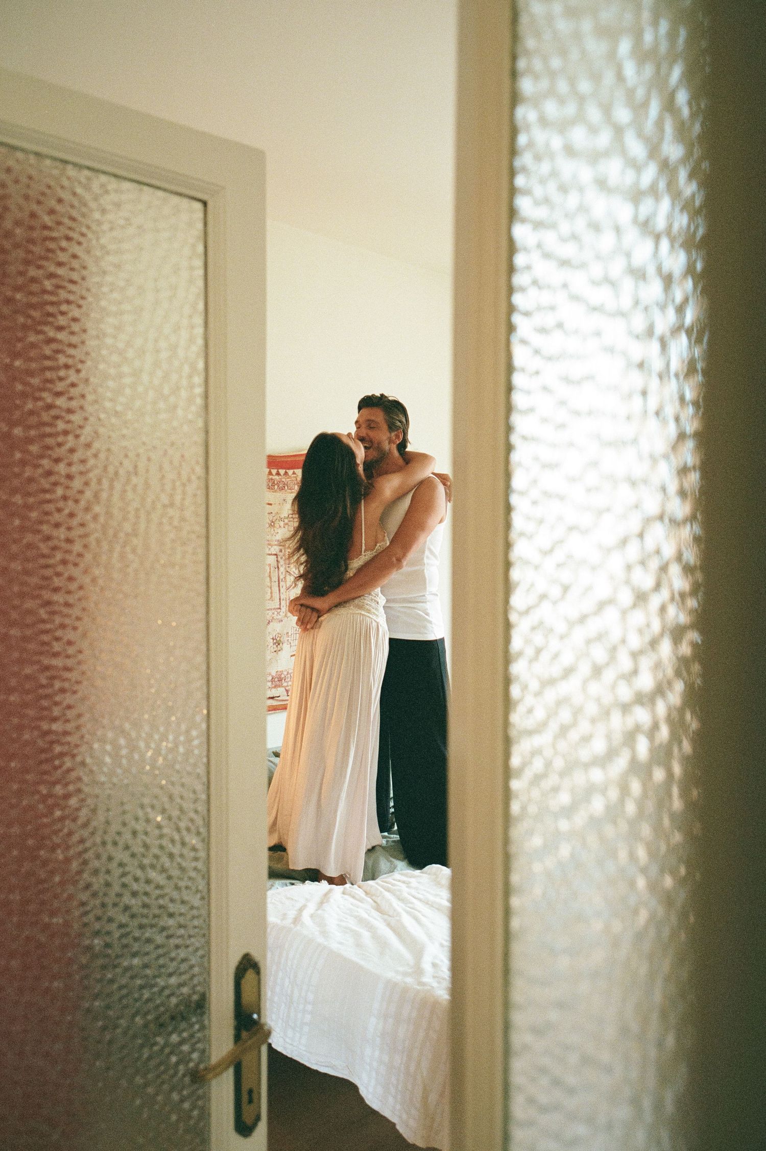 A smiling couple stands closely together on a bed, viewed through a partially open frosted glass bathroom door.