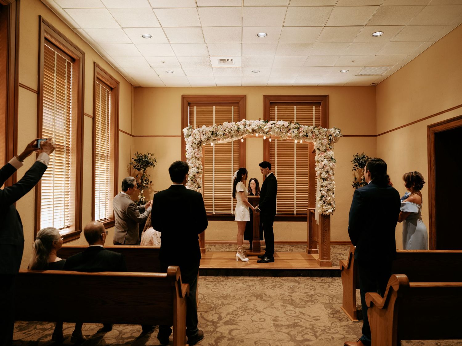 Intimate civil wedding ceremony in small chapel with floral arch, officiant, and small group of witnesses.