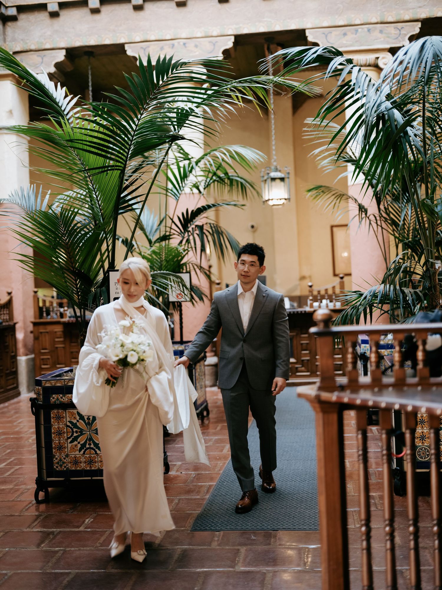 Bride and groom walk through elegant indoor courtyard with lush tropical palm plants.