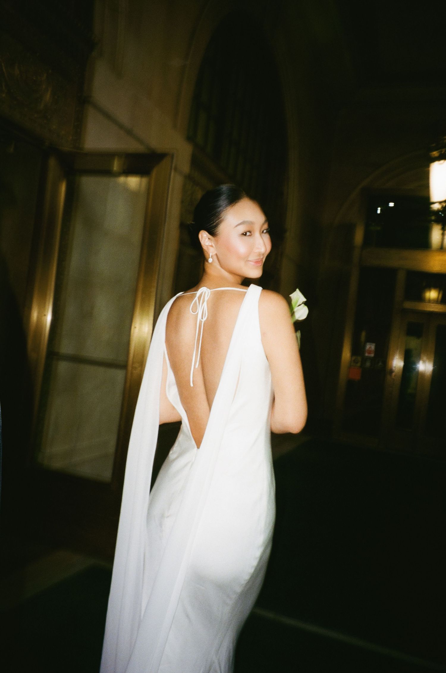 Bride in elegant white backless gown with cape detail turns to smile over shoulder in ornate gold venue.