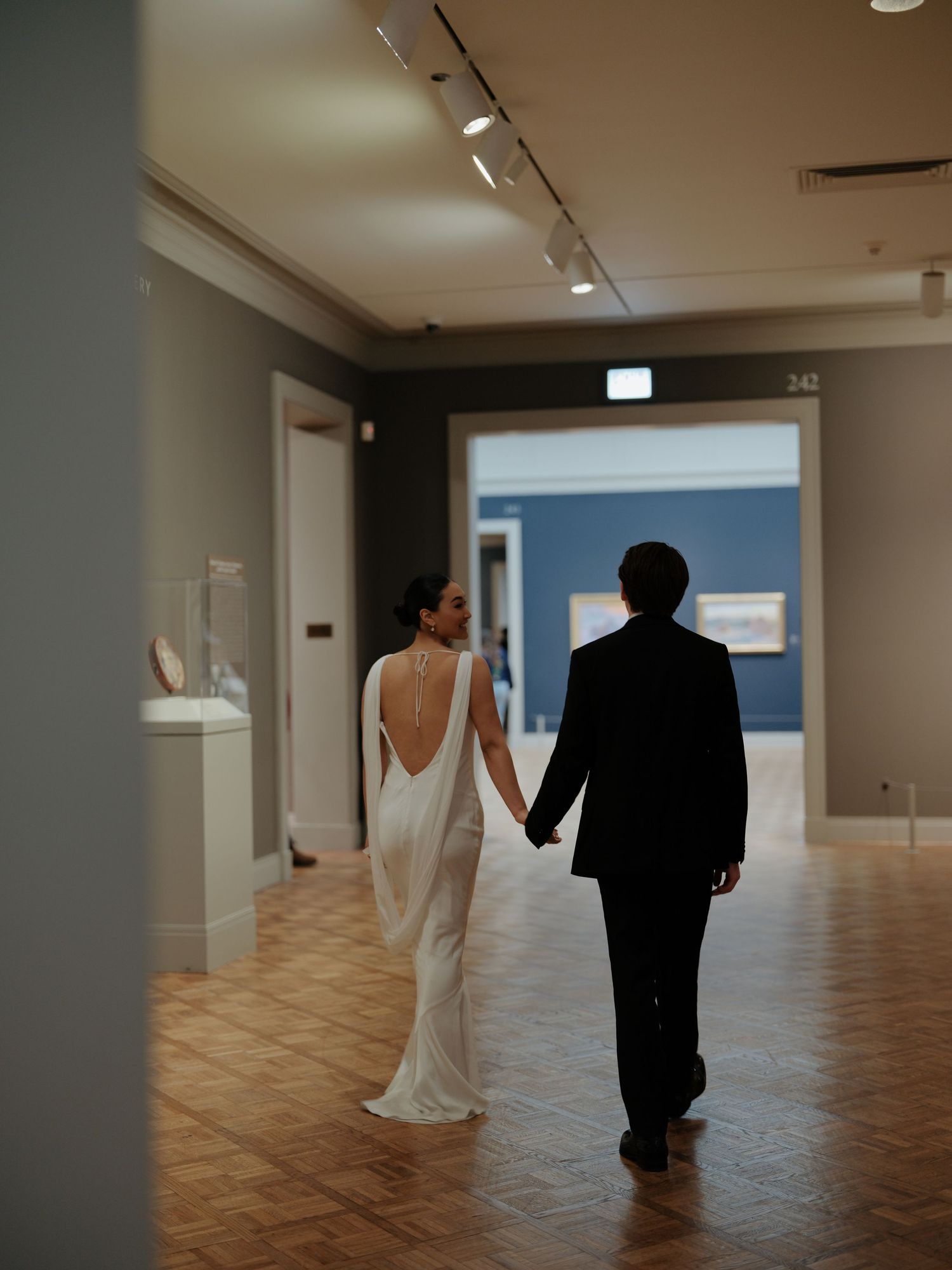 Newlyweds walking hand in hand through an art museum gallery, bride in elegant backless white gown.