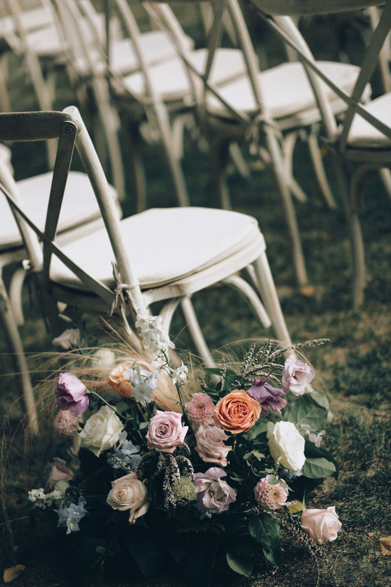 Colorful wedding bouquet of pink, peach, and white roses resting on grass beside rustic wooden ceremony chairs.