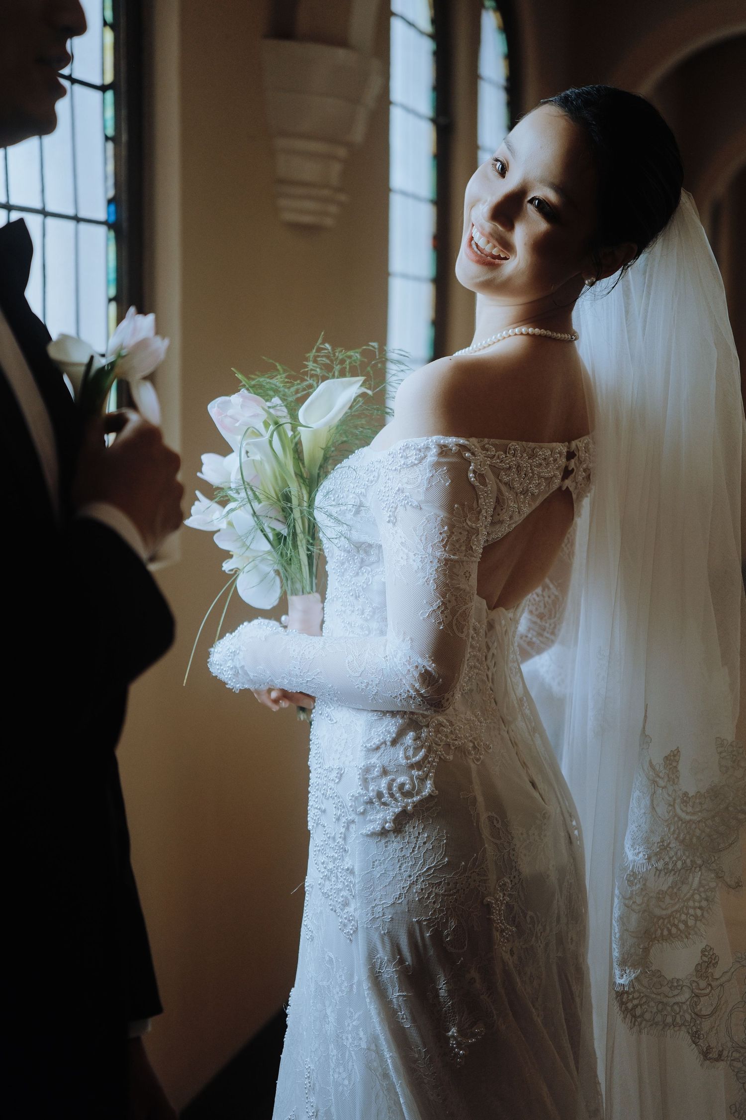 Smiling bride in off-shoulder lace gown with veil holds white tulip bouquet in church.