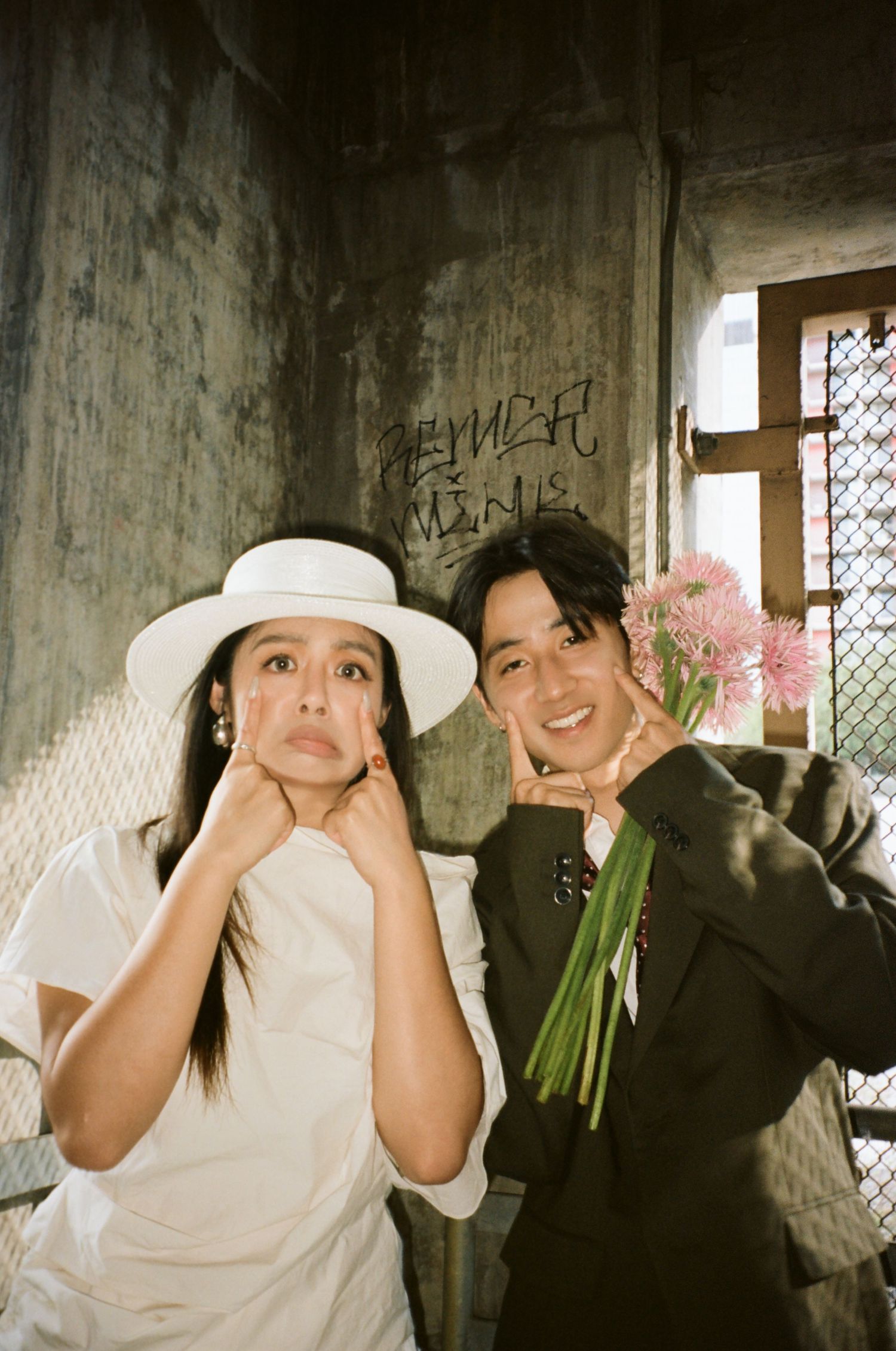 Playful couple poses in rustic interior, woman in white hat and dress, man in dark suit holds pink gerbera flowers.