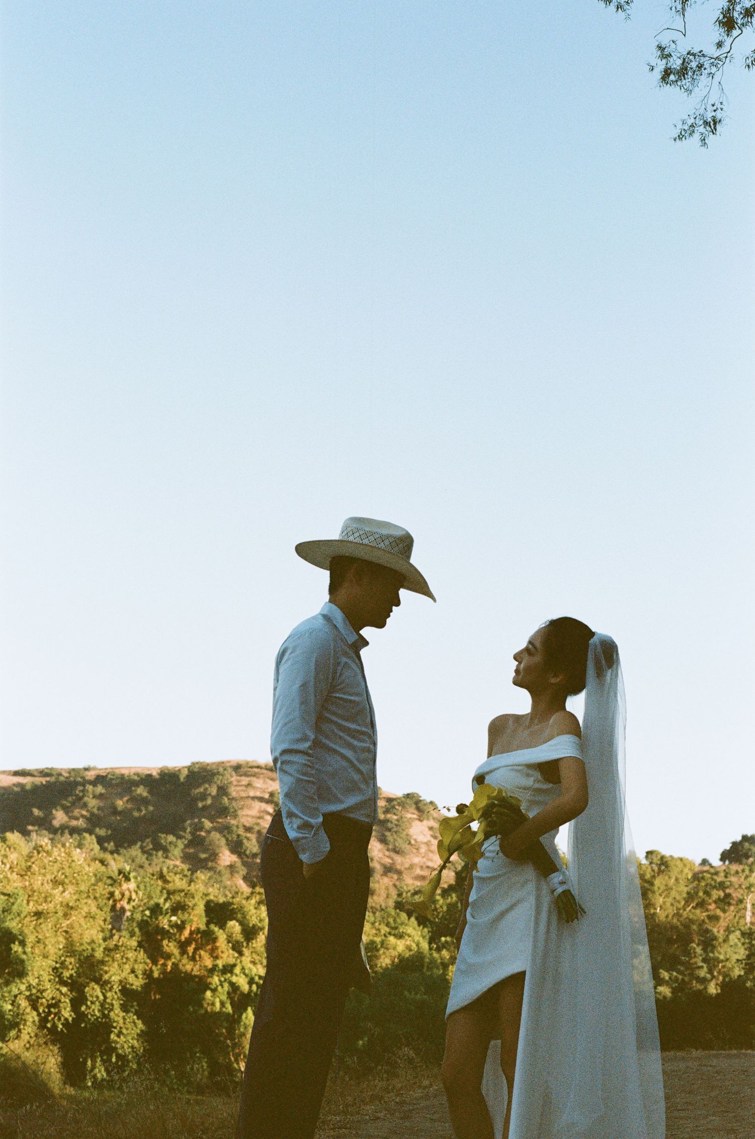 Groom in cowboy hat and bride in off-shoulder gown face each other in silhouette against a clear blue sky outdoors.