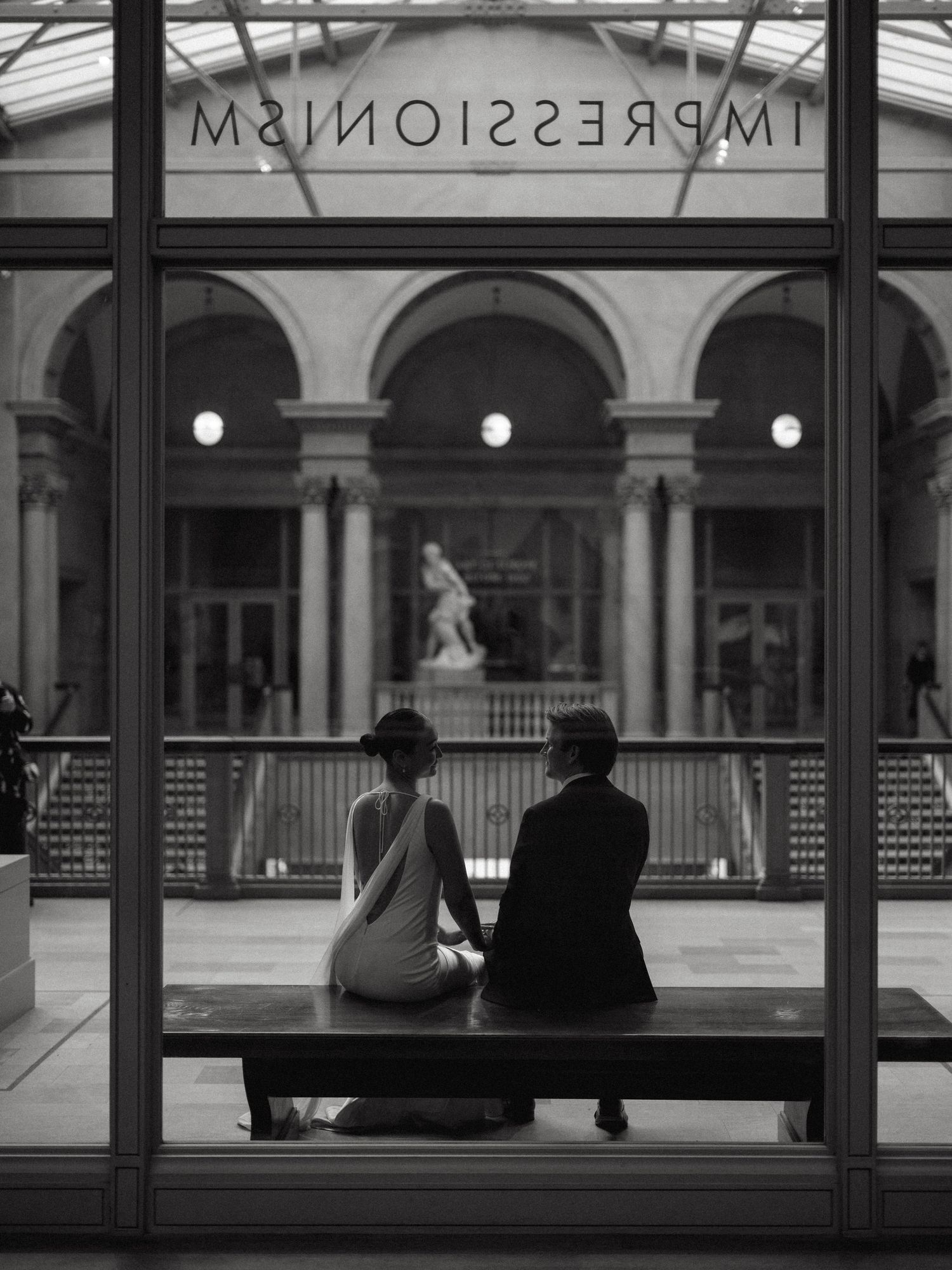 Black and white photo of newlyweds sitting on a bench beneath an Impressionism sign in a grand museum.