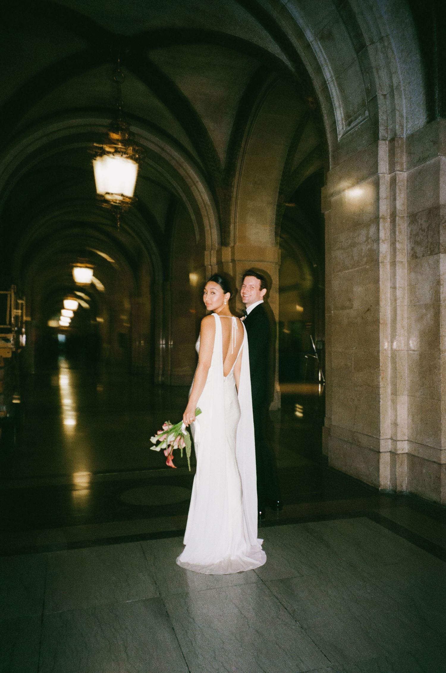 Couple poses in stunning arched stone corridor, bride in backless white gown holds bouquet, groom in tuxedo.