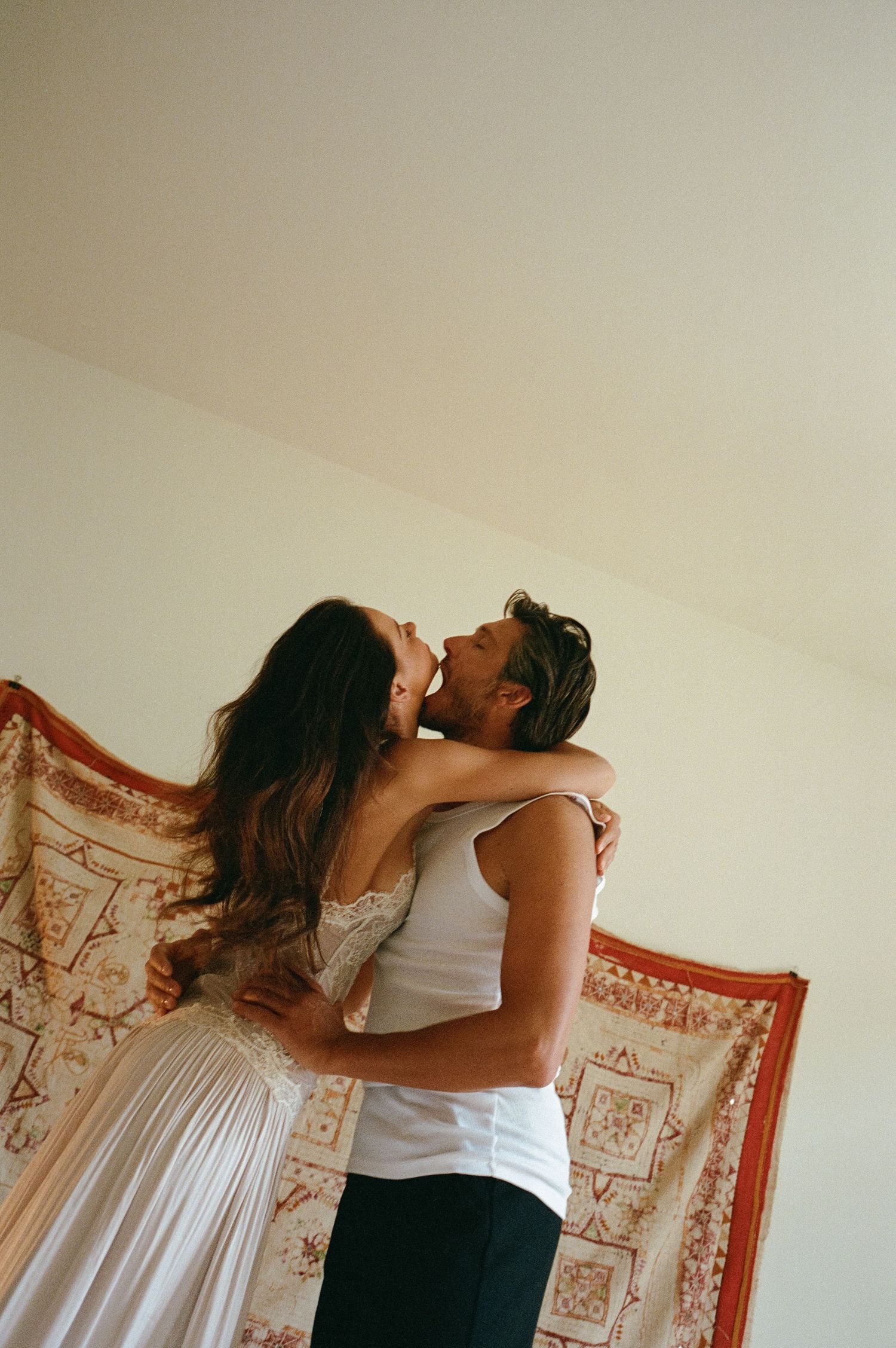 Romantic couple embracing and kissing in a cozy room with a decorative tapestry backdrop.
