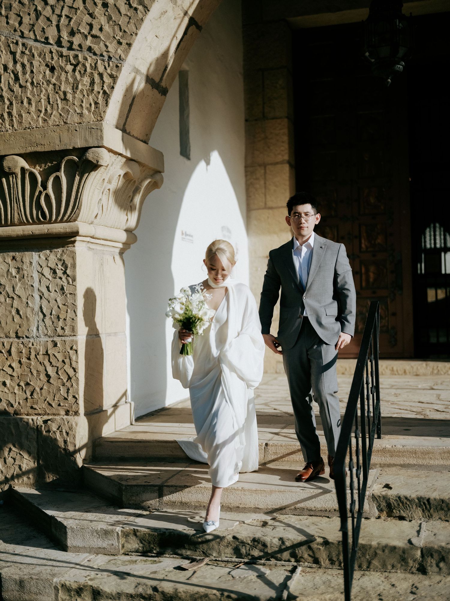 Bride and groom descend sunlit stone steps of a historic Spanish-style building.