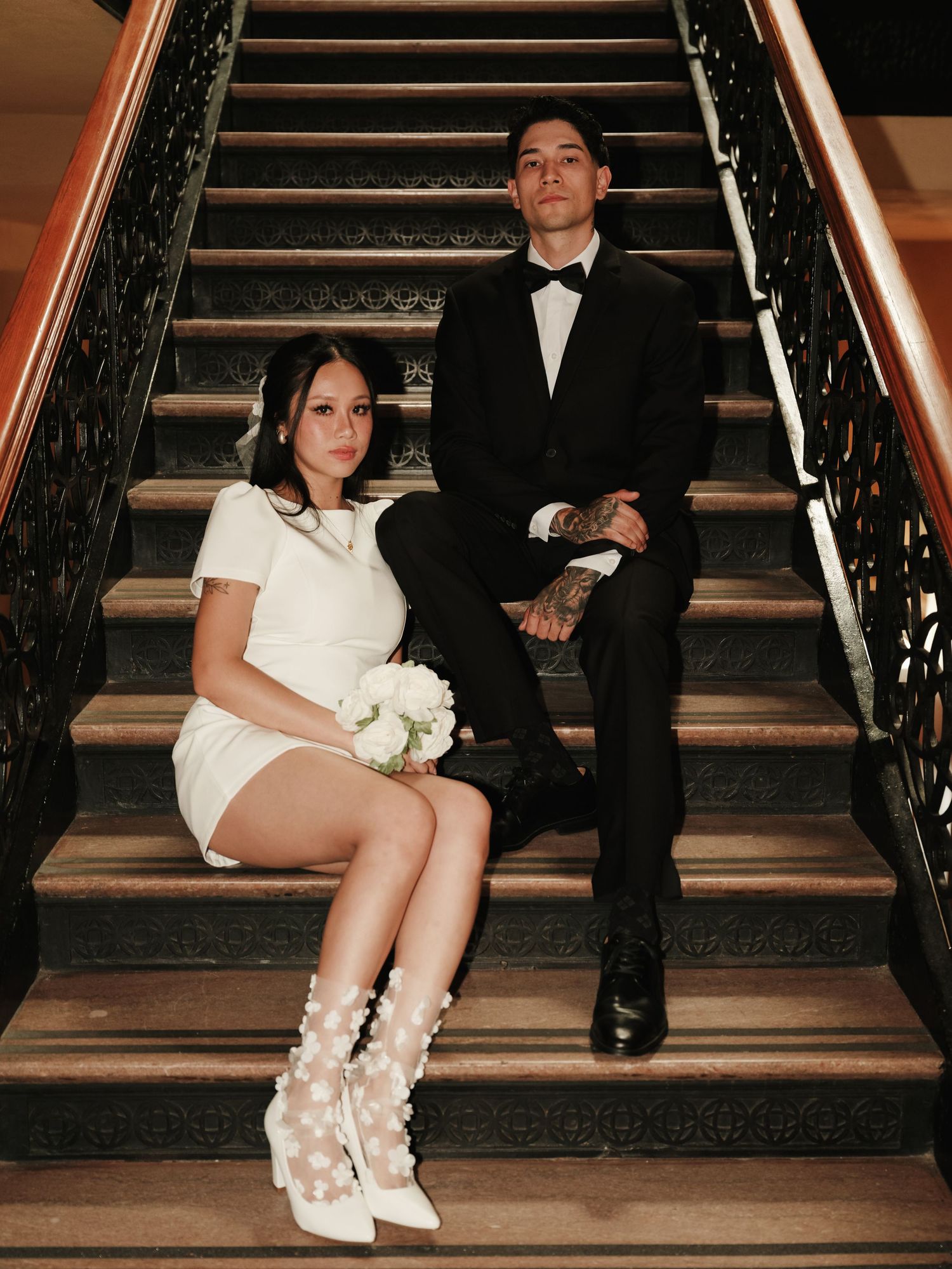 Stylish newlyweds pose on ornate staircase, bride wearing floral white boots and mini dress.