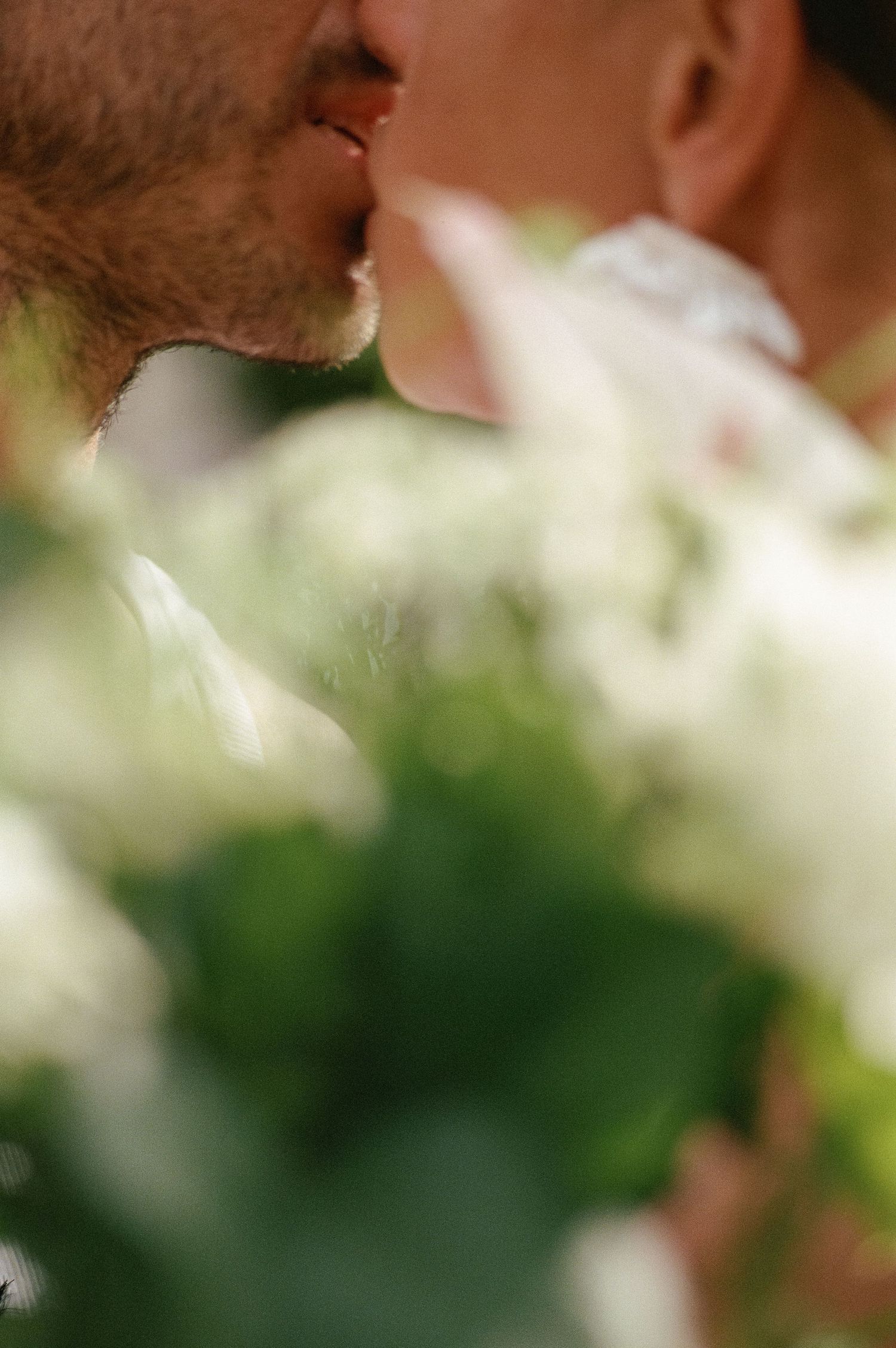 Close-up of a couple kissing behind a blurred white wedding bouquet in soft natural light.