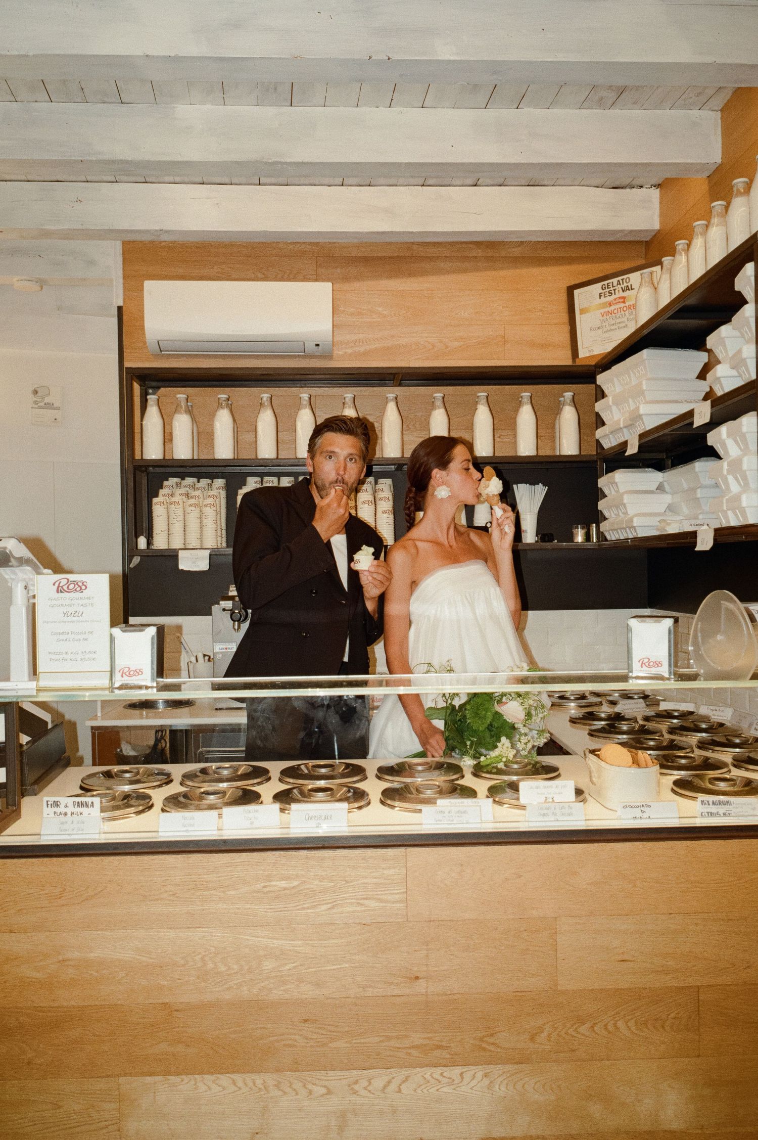 Bride and groom enjoying gelato together at an Italian gelateria counter on their wedding day.