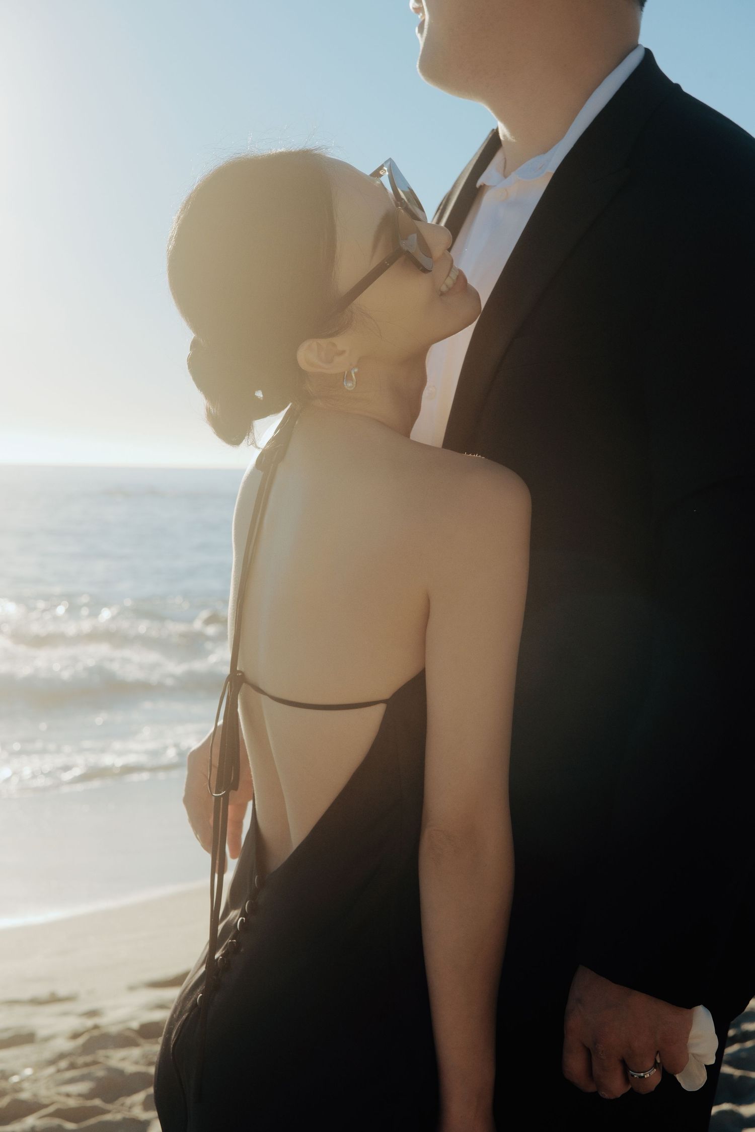 Bride in backless black dress leaning on groom in suit at beach.