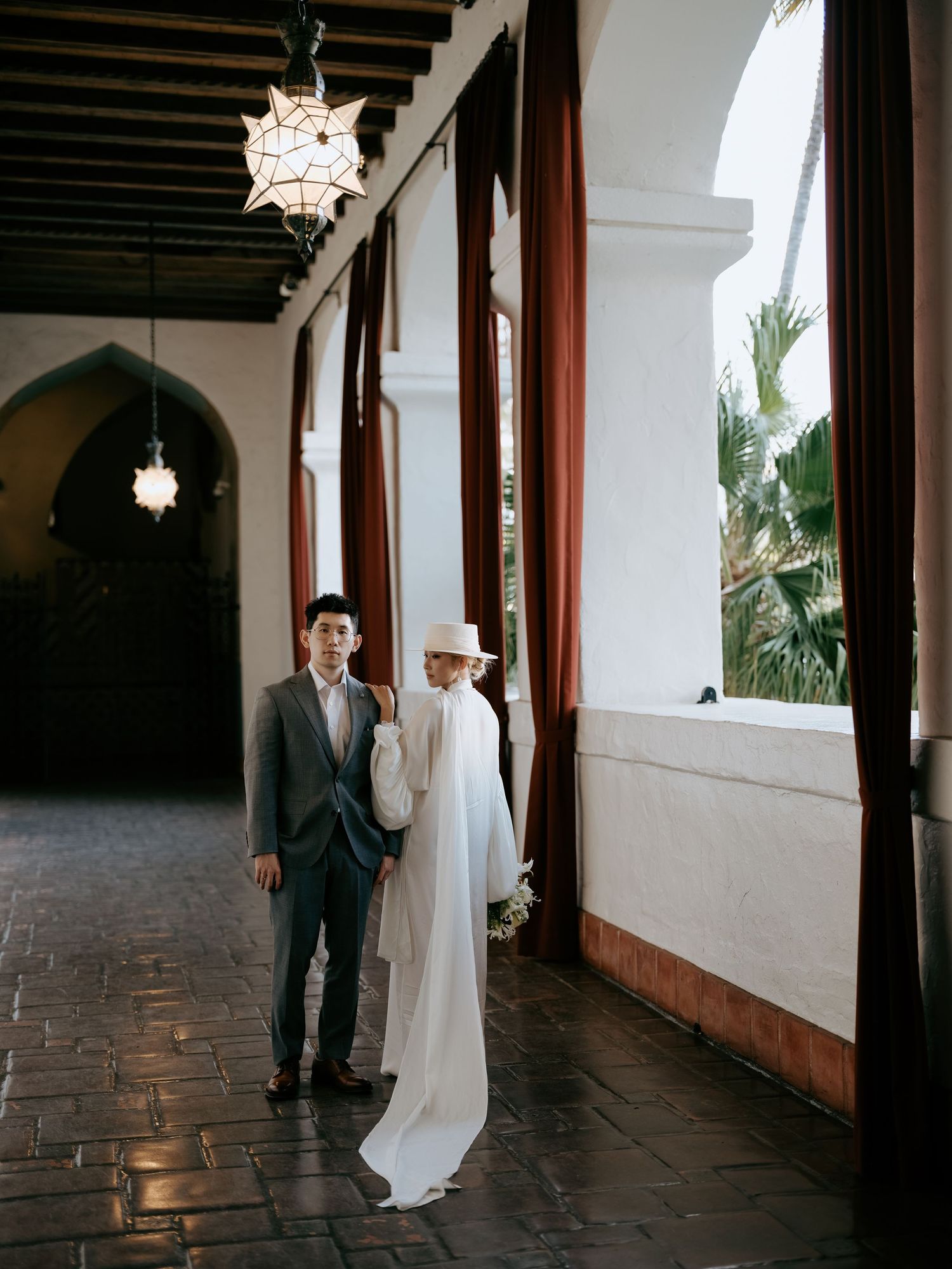 Bride in white gown and hat with groom in gray suit posing in elegant arched corridor with ornate lanterns.
