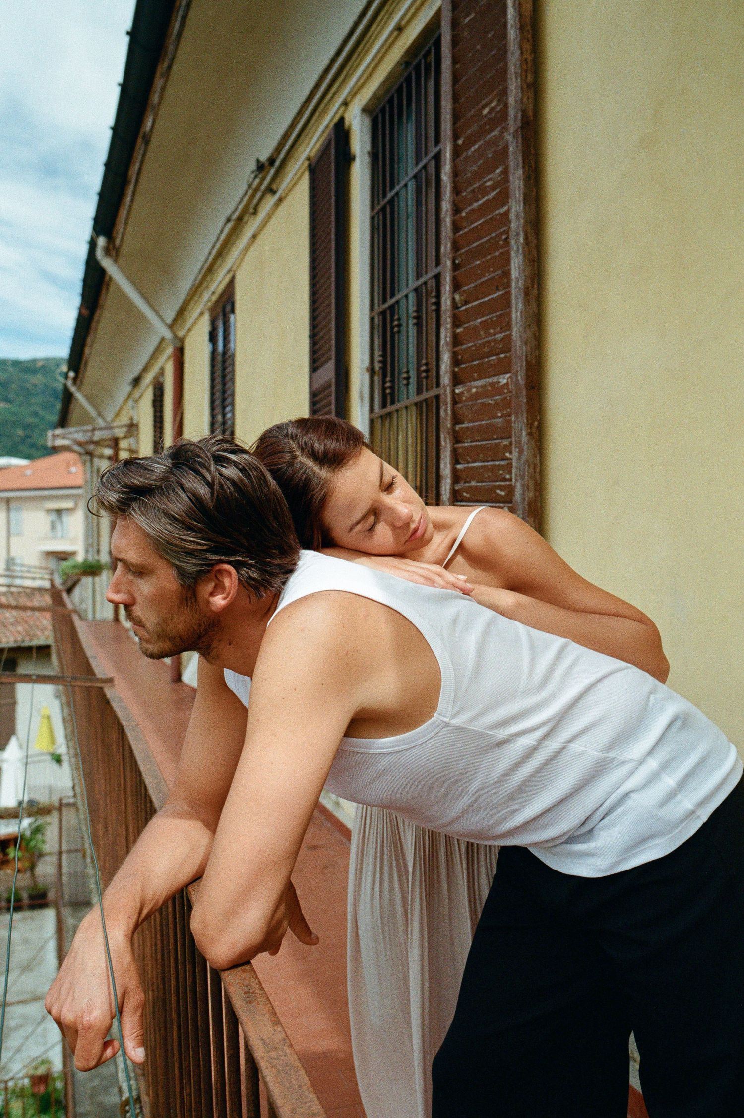 Couple in casual attire leans on a wooden balcony railing of a European building on a cloudy day.