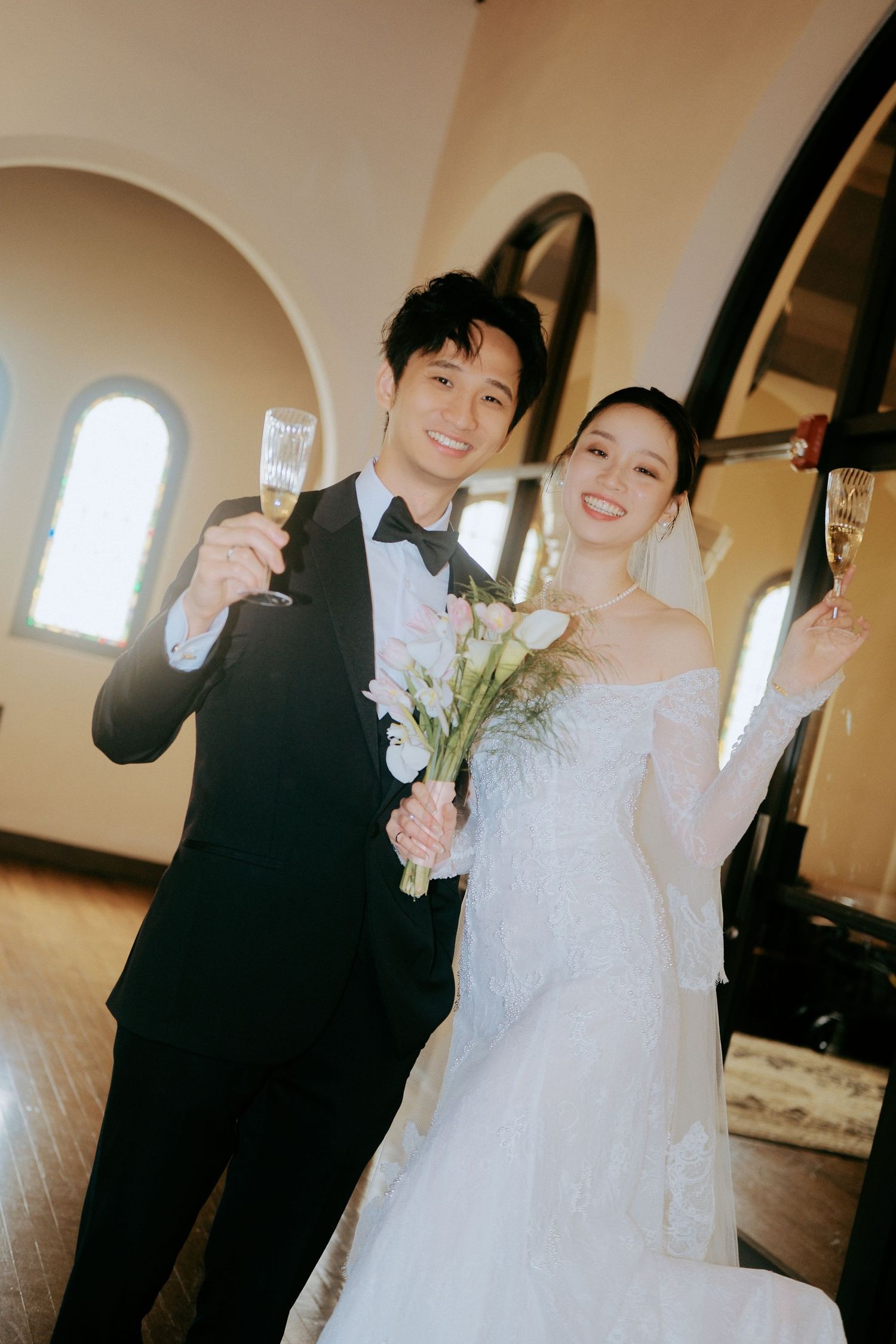 Happy couple raising champagne glasses in a toast inside a sunlit chapel.