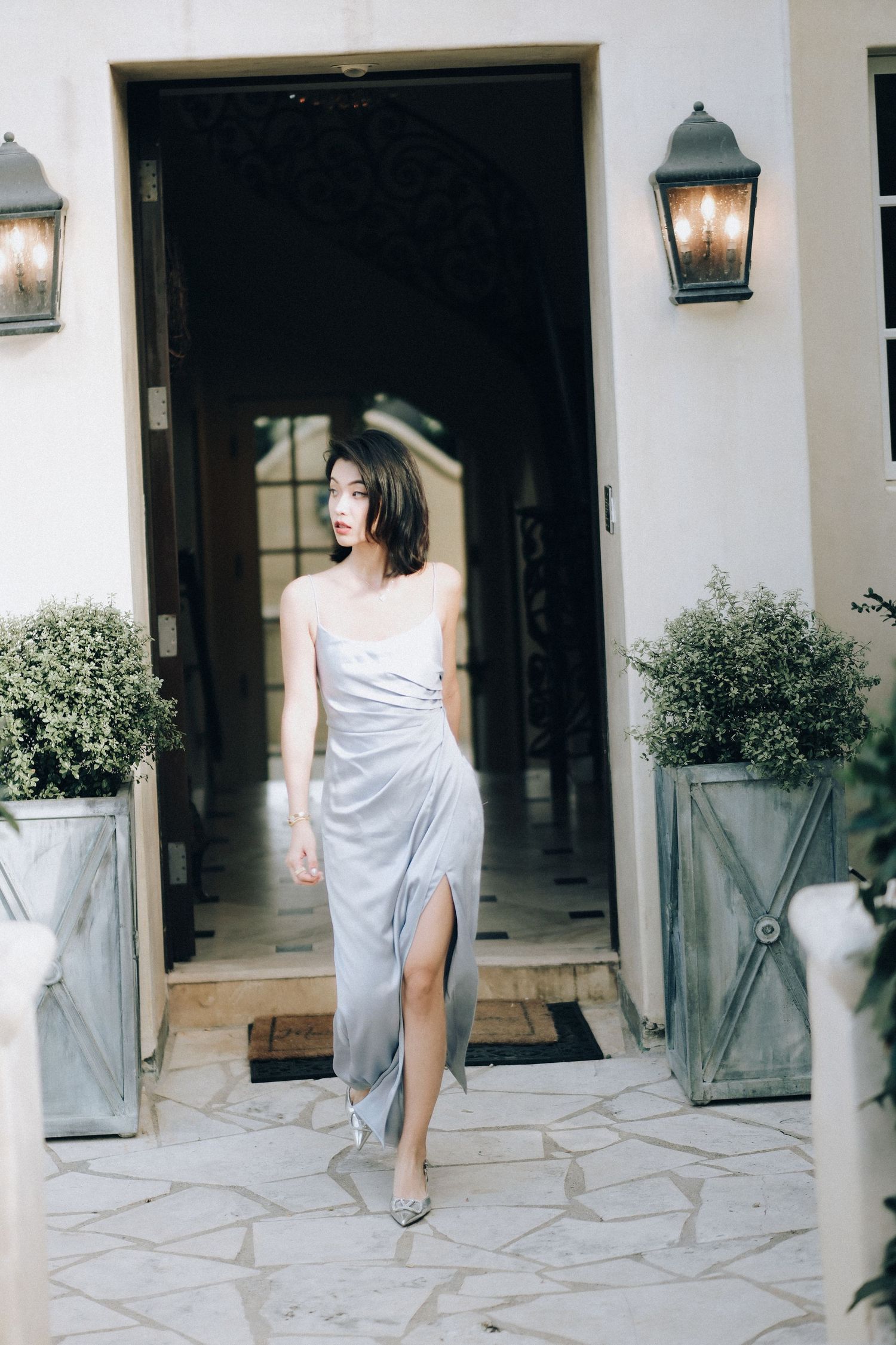 Woman in silver slit dress walks through grand doorway flanked by lanterns and potted plants.