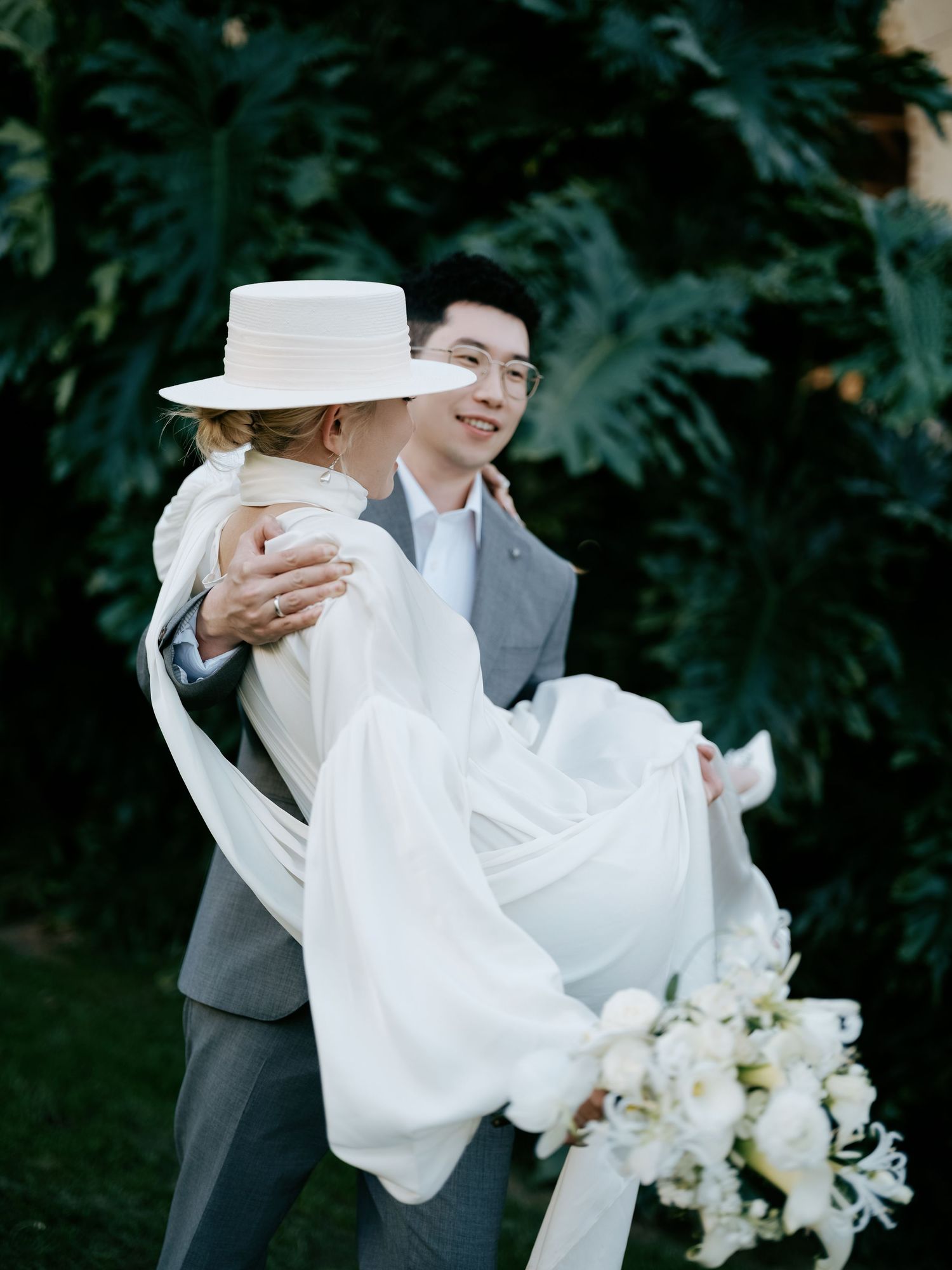Groom in gray suit carries bride in white cape dress and wide-brim hat among lush greenery.