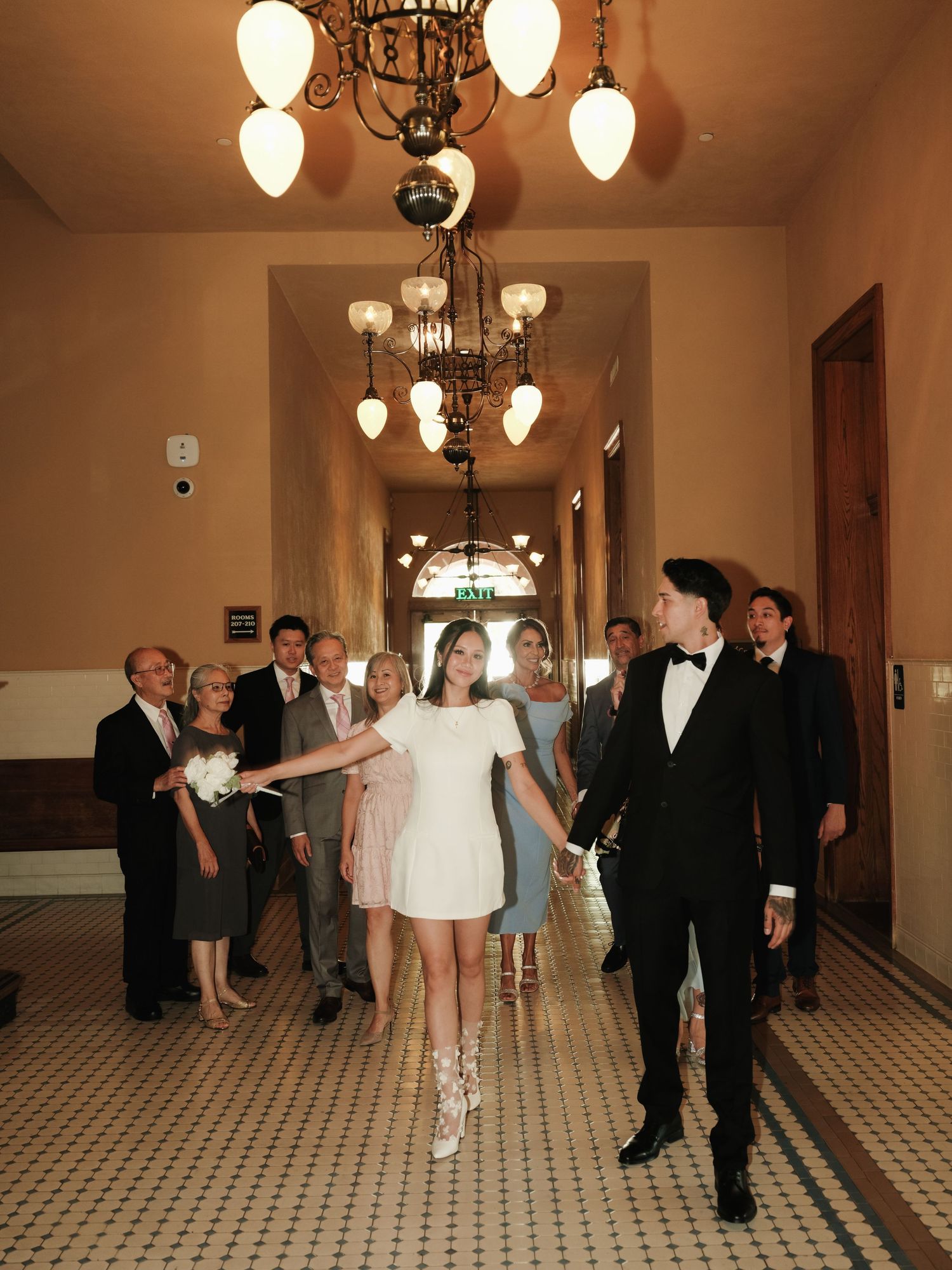 Bride and groom walk through grand corridor with family following, bride reaching back to hold hands.