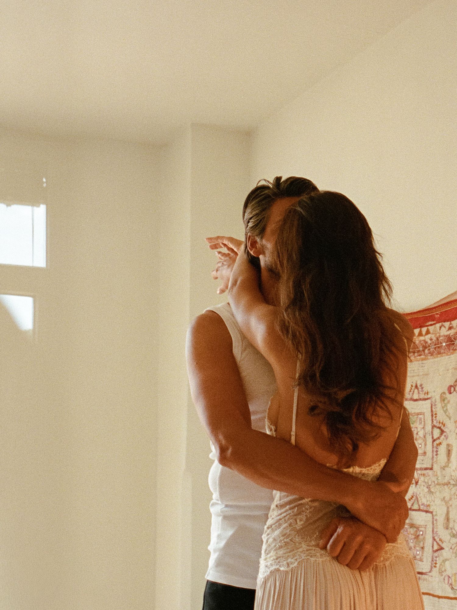 A couple shares a tender embrace and kiss near a bright window with a colorful tapestry visible in the background.
