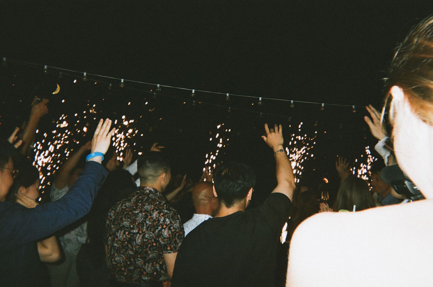 Wedding guests celebrating at night, raising hands amid sparkling fireworks and string lights.