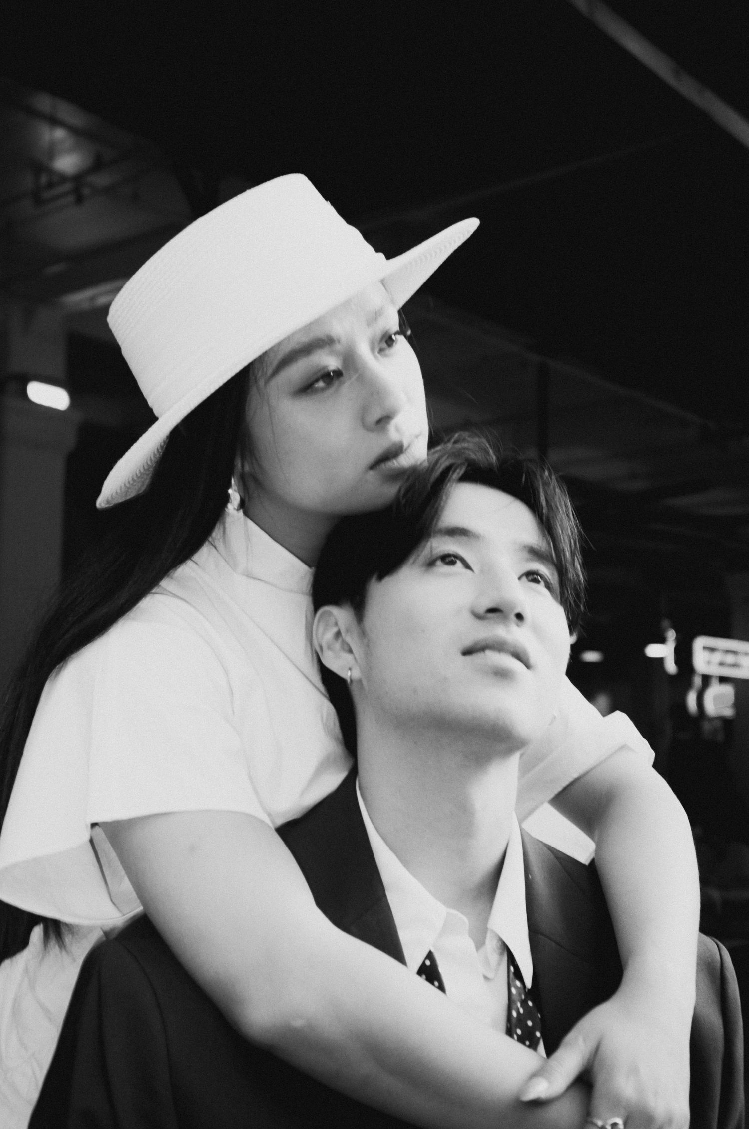 Black and white portrait of couple, woman in white hat embraces man from behind in intimate pose.