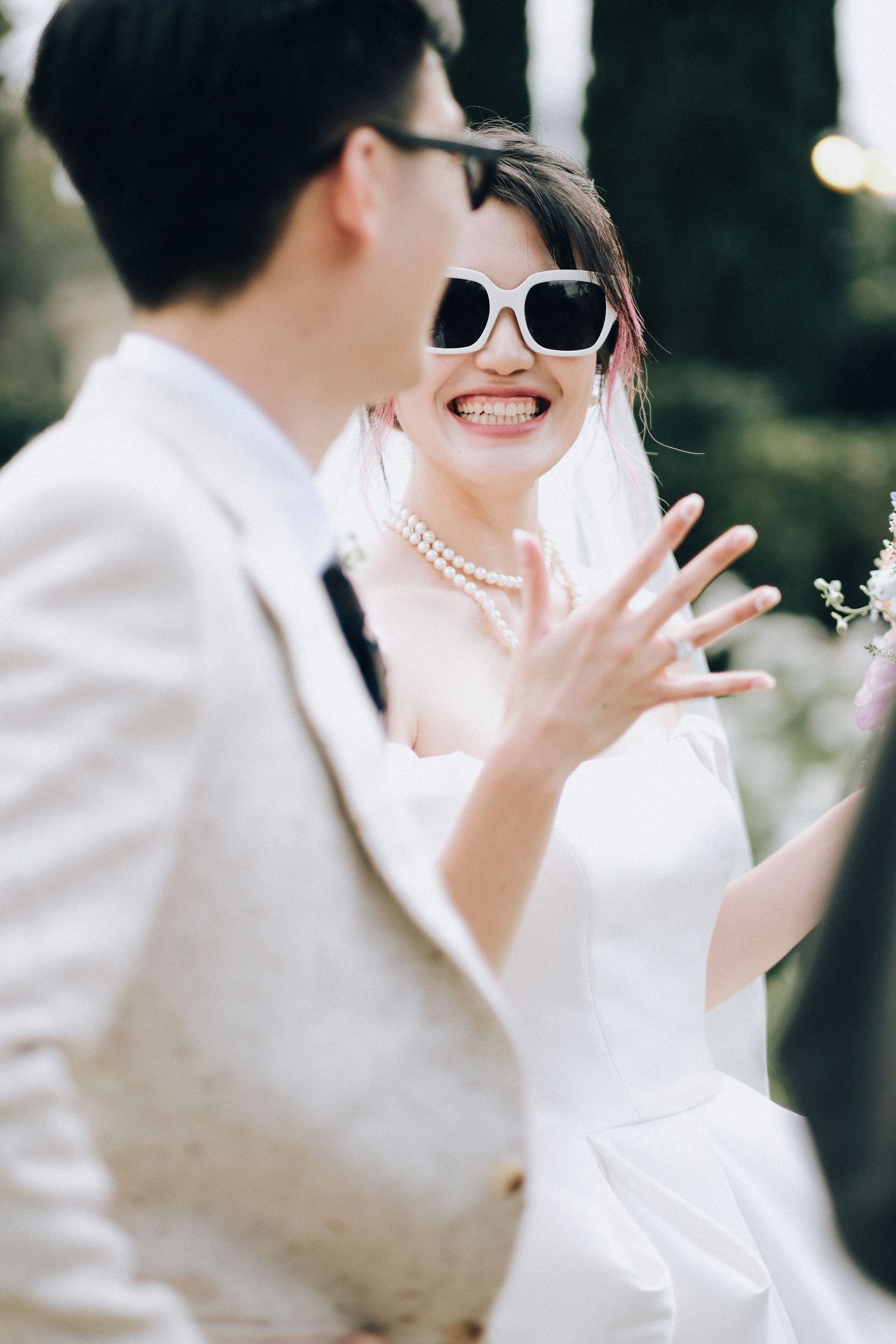 Smiling bride in white sunglasses and pearl necklace shows off ring while talking to groom.