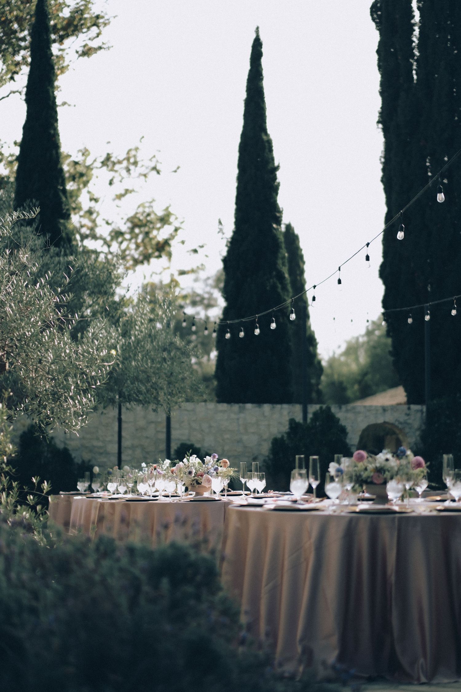 Romantic outdoor wedding reception long table set with blush linens and florals beneath cypress trees and string lights.