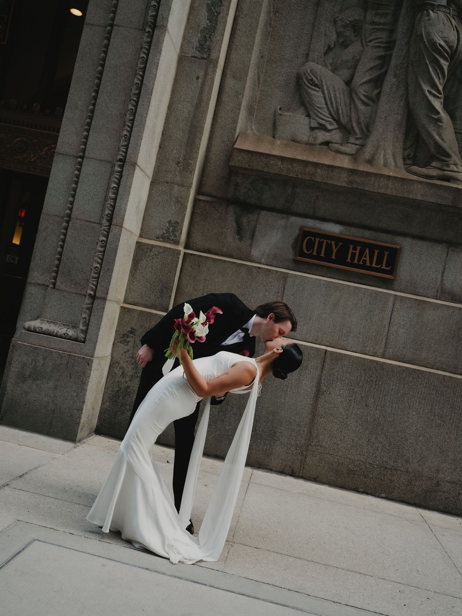Groom dipping bride in a dramatic kiss outside City Hall, bride in flowing white gown with cape detail.