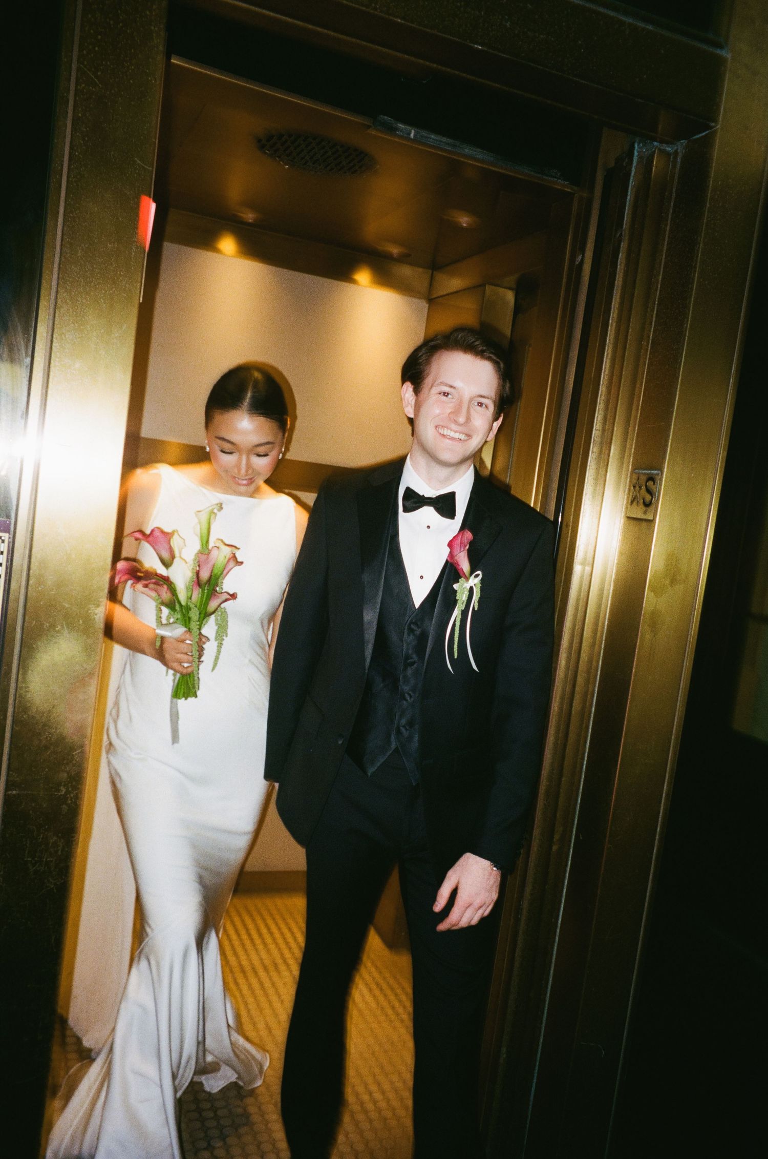 Groom in black tuxedo leads bride in white gown out of a gold elevator, holding red bouquet.