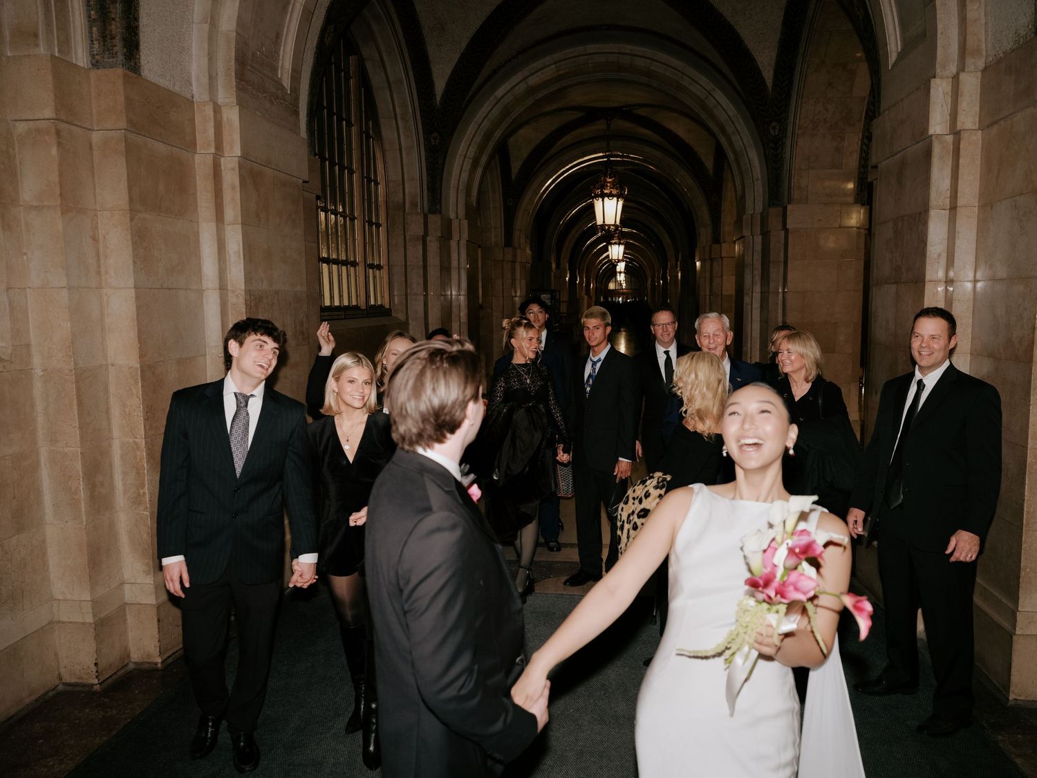 Bride and groom holding hands leading wedding party through ornate stone archway corridor.