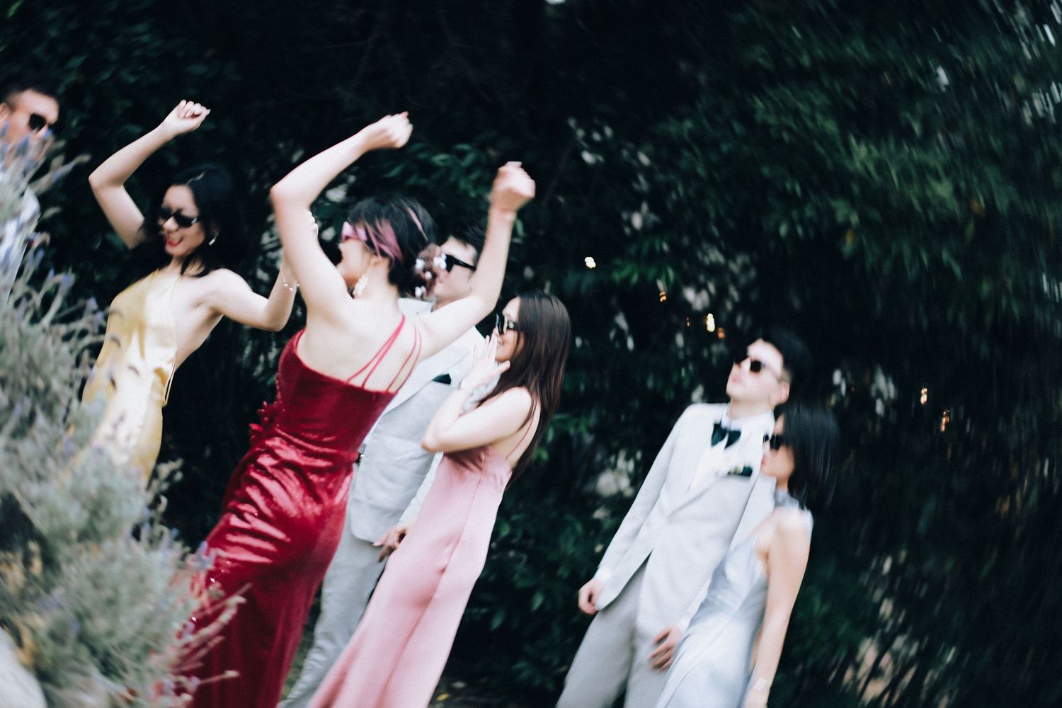 Wedding guests in colorful formal attire dancing outdoors among lush green trees at reception celebration.