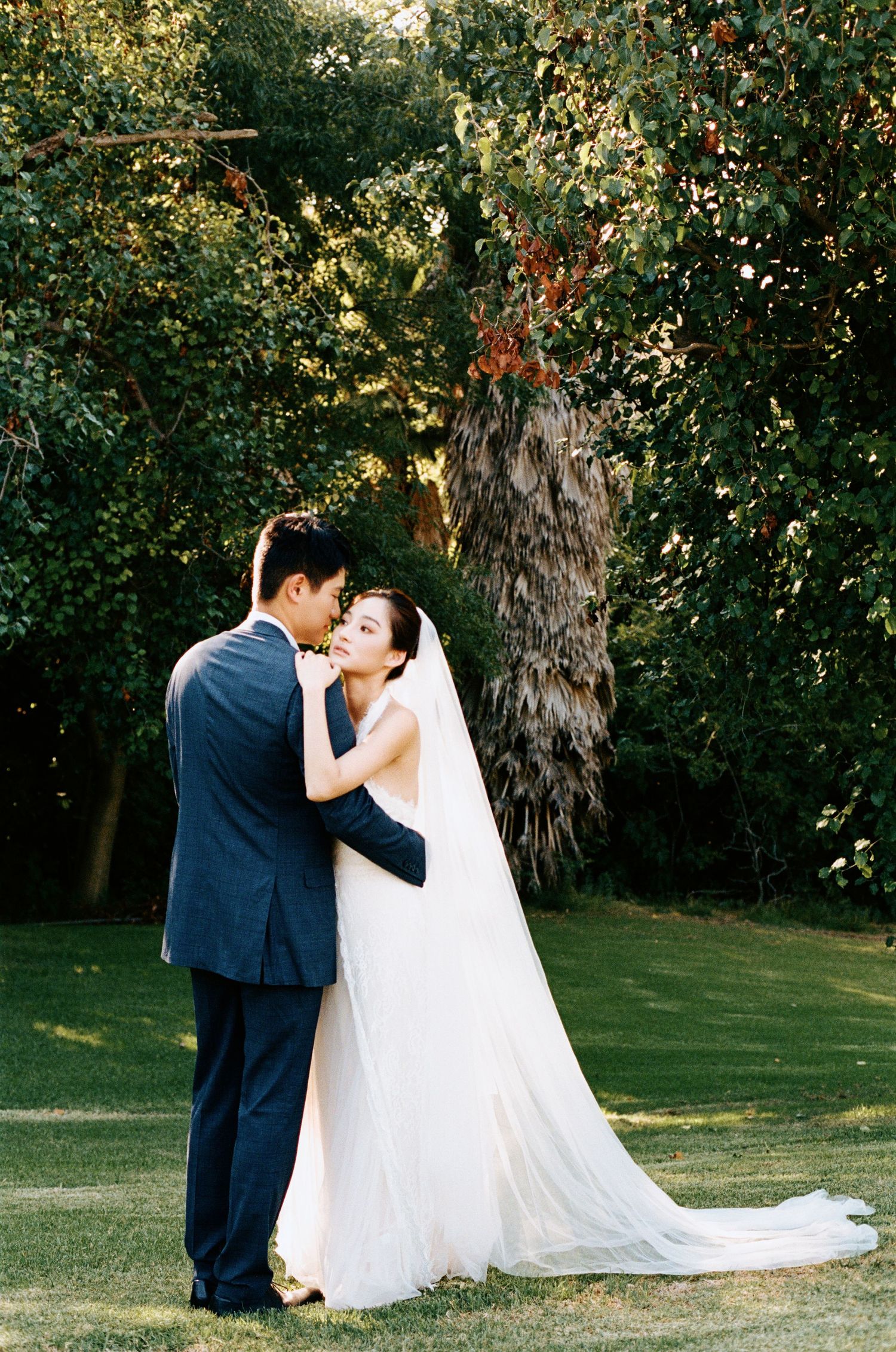 Bride and groom embrace on a lush green lawn surrounded by trees in golden hour light.