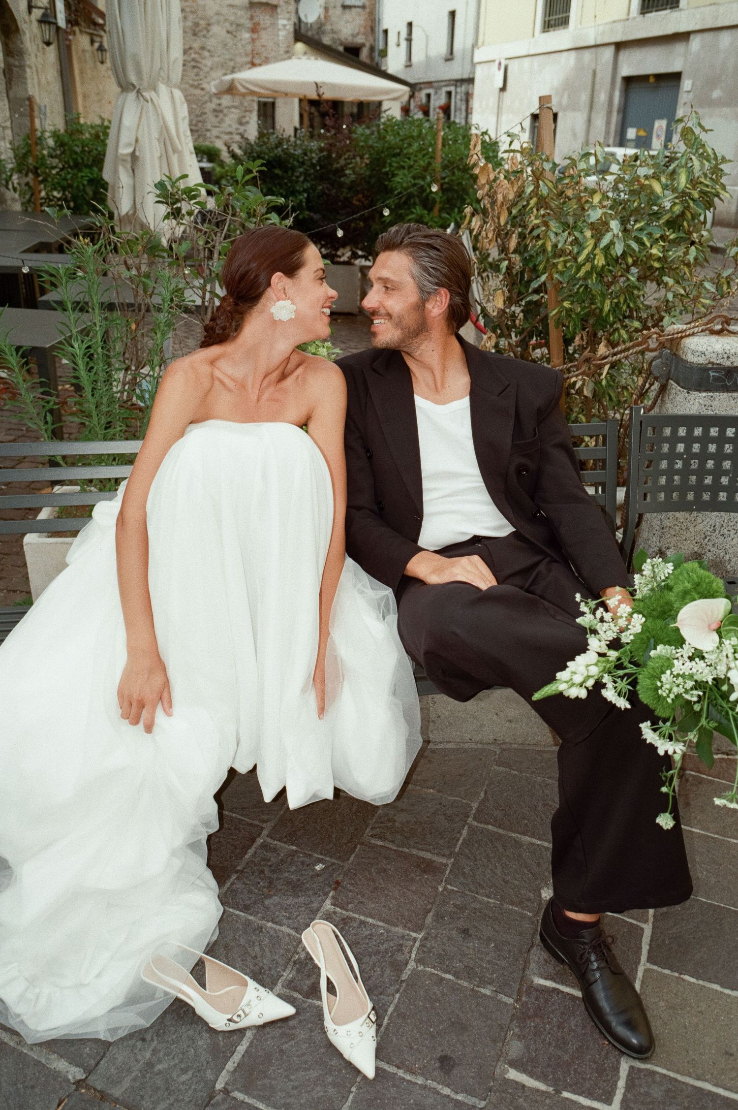 Smiling bride and groom seated on a bench in an Italian piazza with white floral bouquet.