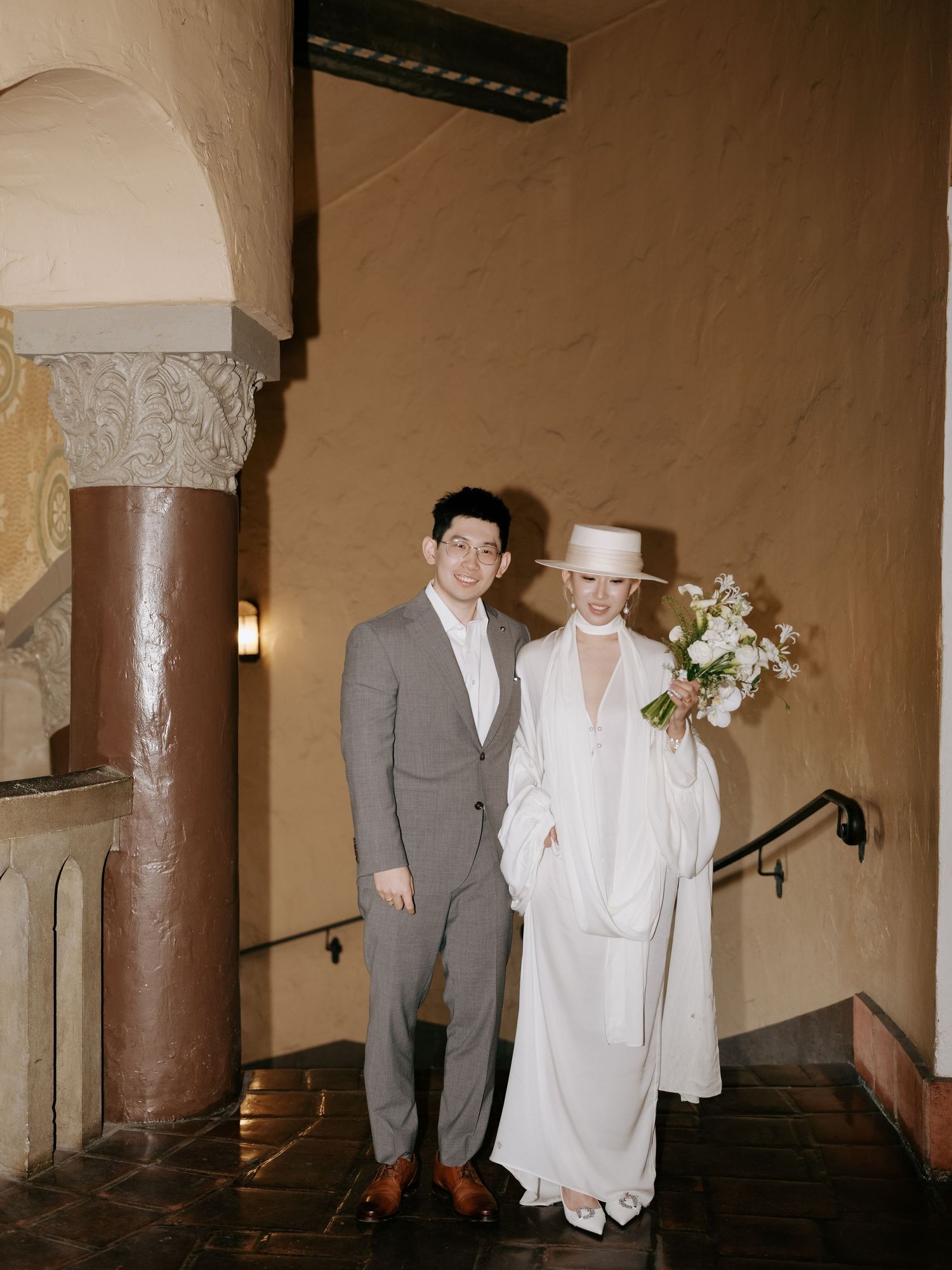Wedding couple posing on stairs, bride in white cape dress and wide-brim hat holding floral bouquet.