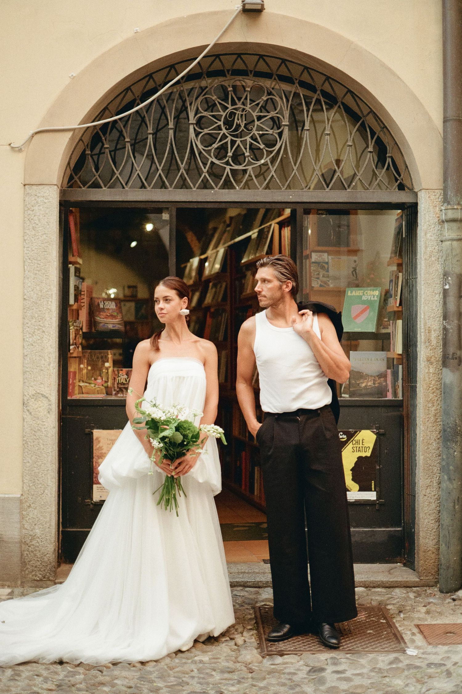 Bride in white strapless gown holds bouquet beside groom in white tank top outside Italian shop.