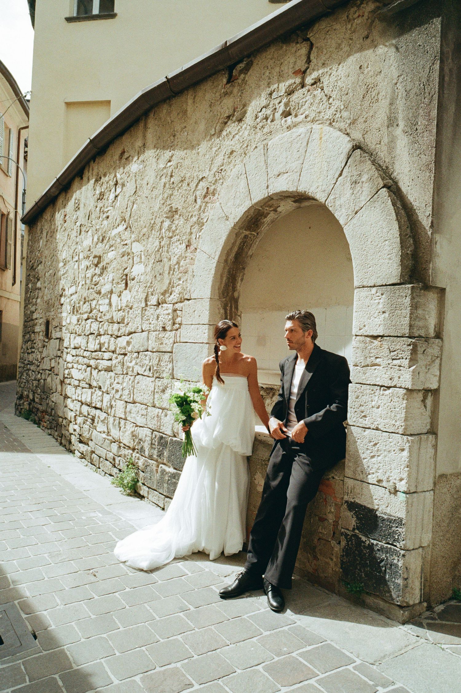 Bride in white gown and groom in black suit pose by a stone arch wall in a European cobblestone alley.