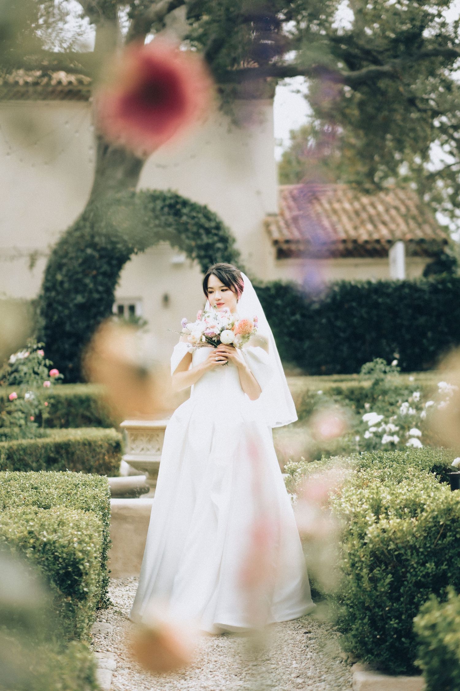 Bride in white off-shoulder gown holds bouquet while standing in a manicured garden pathway.