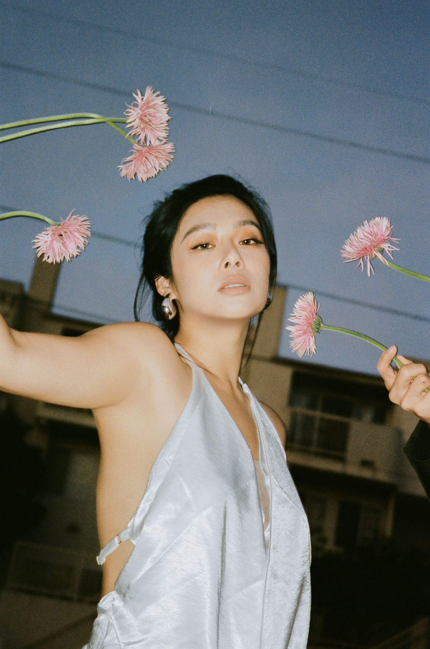 Woman in silver halter dress holds pink gerbera flowers against twilight sky in artistic outdoor portrait.